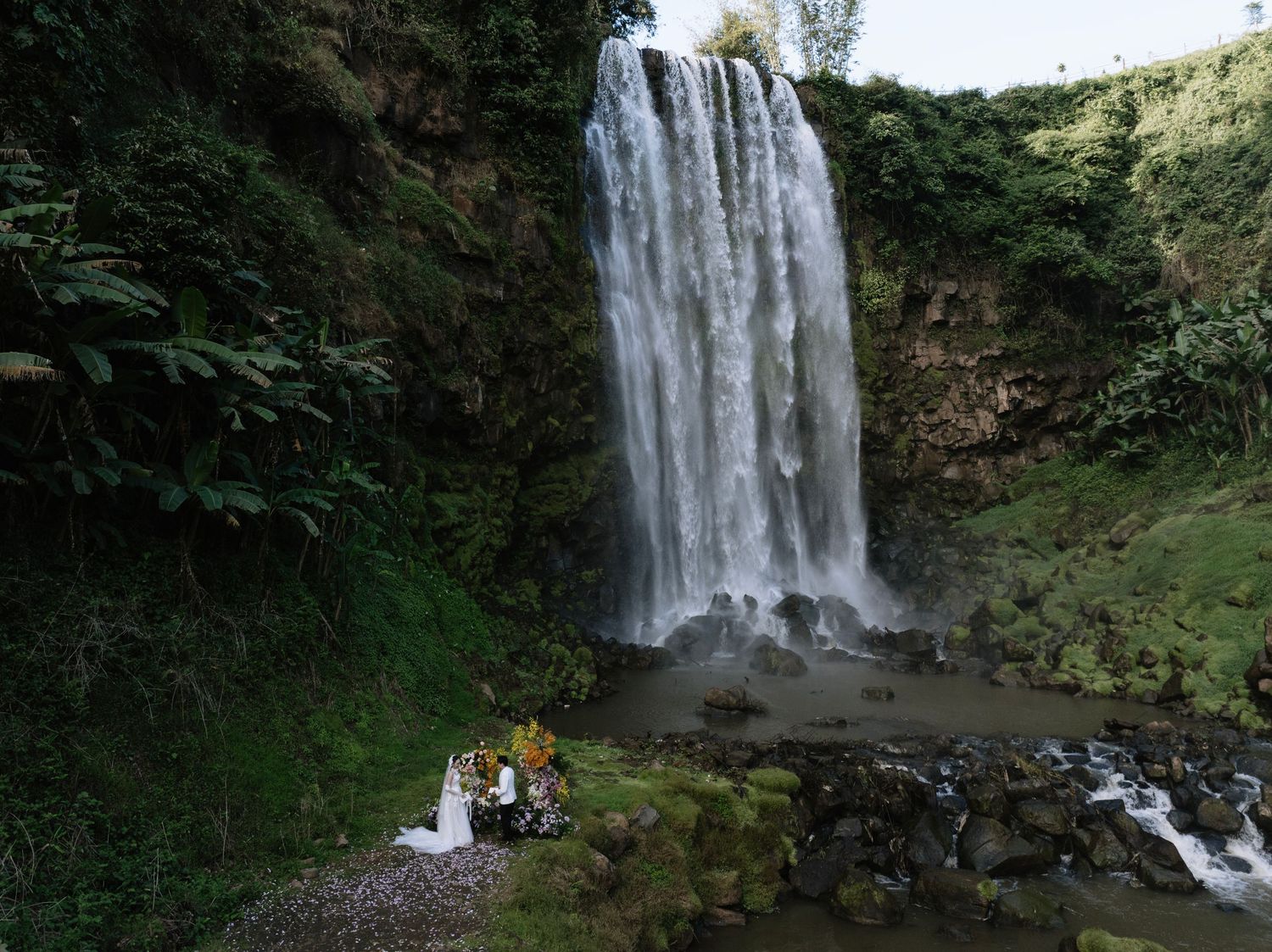 Couple poses before a stunning tall waterfall surrounded by lush green tropical vegetation.