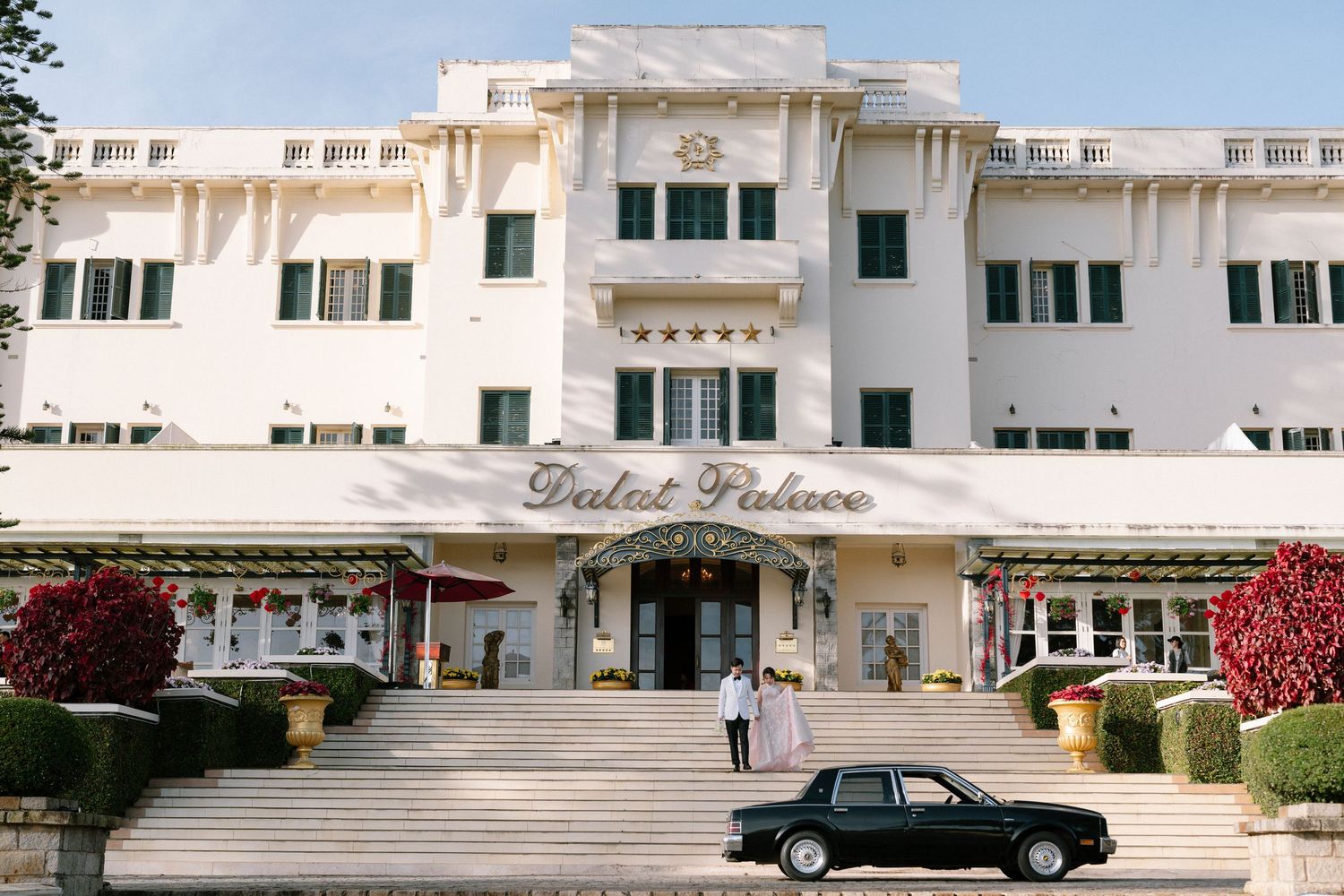 Couple stands before the grand white Dalat Palace Hotel entrance with a vintage black car.