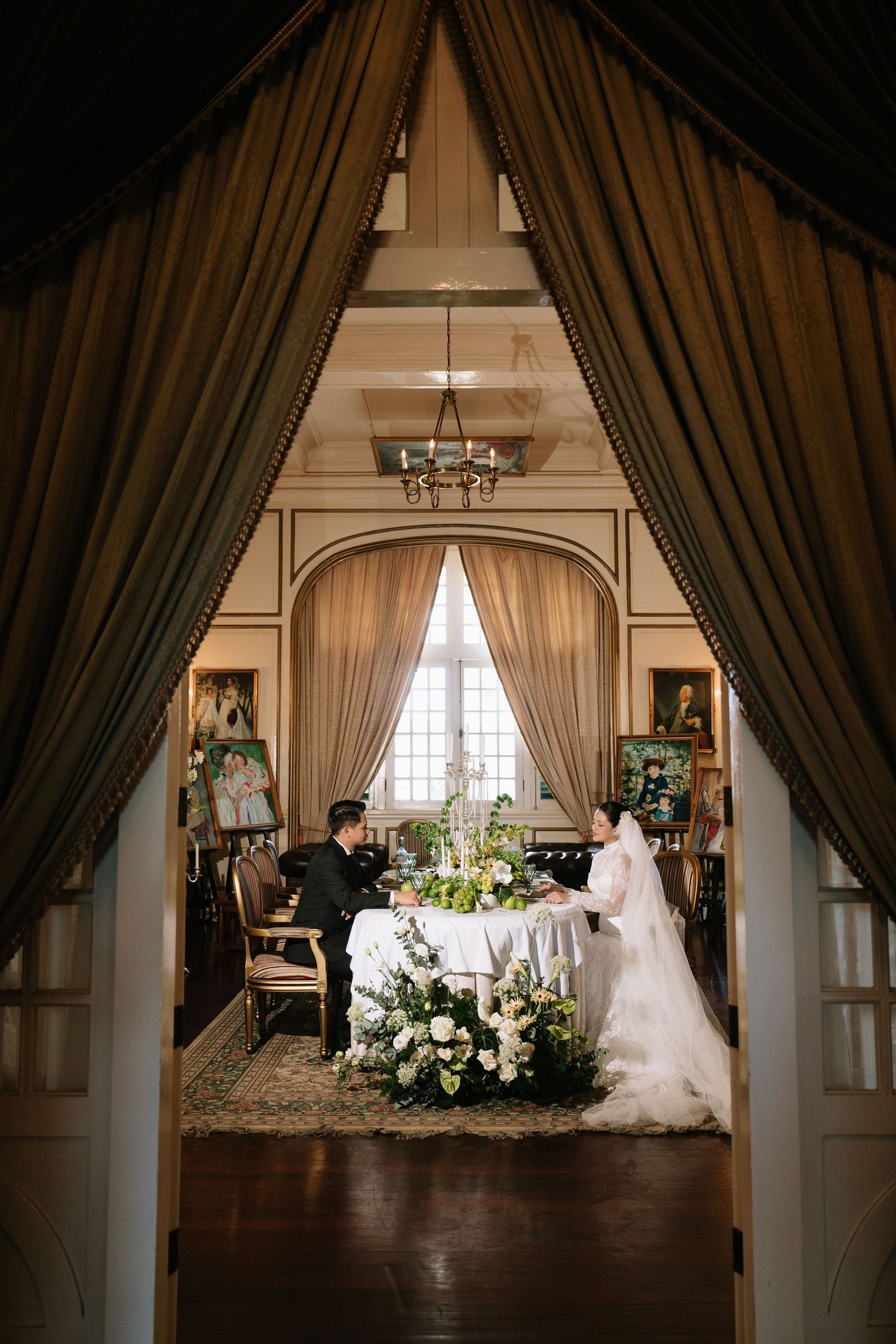 Bride and groom sit at a floral table framed by elegant draped curtains in a classic interior.