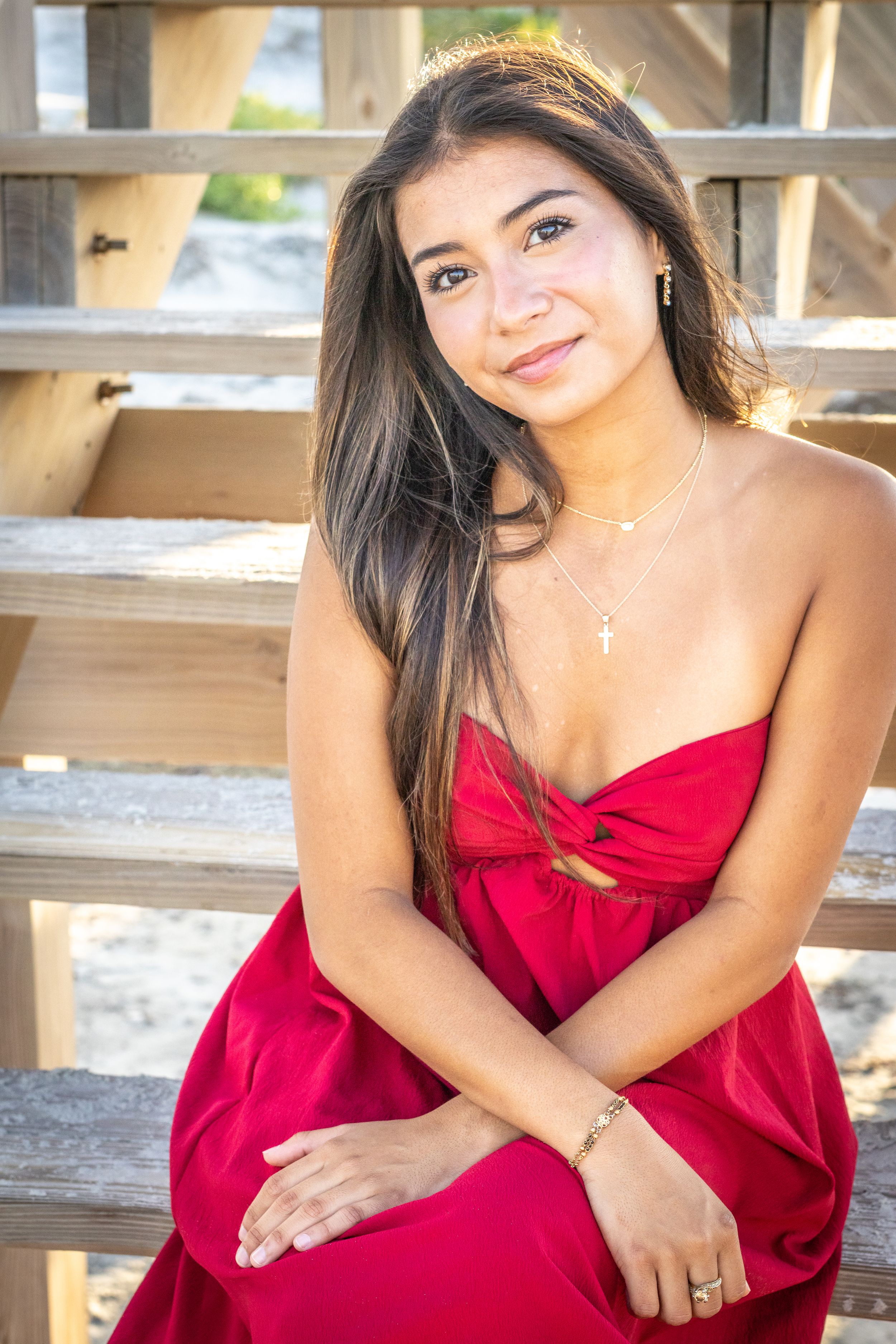 Young woman in red strapless dress with cross necklace sits on wooden steps, smiling softly at camera.