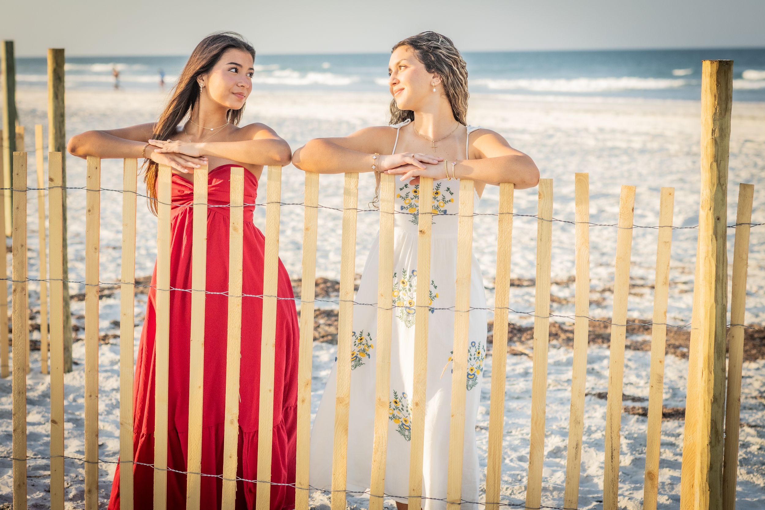 Two women in elegant maxi dresses pose by a wooden beach fence with ocean waves in the background.