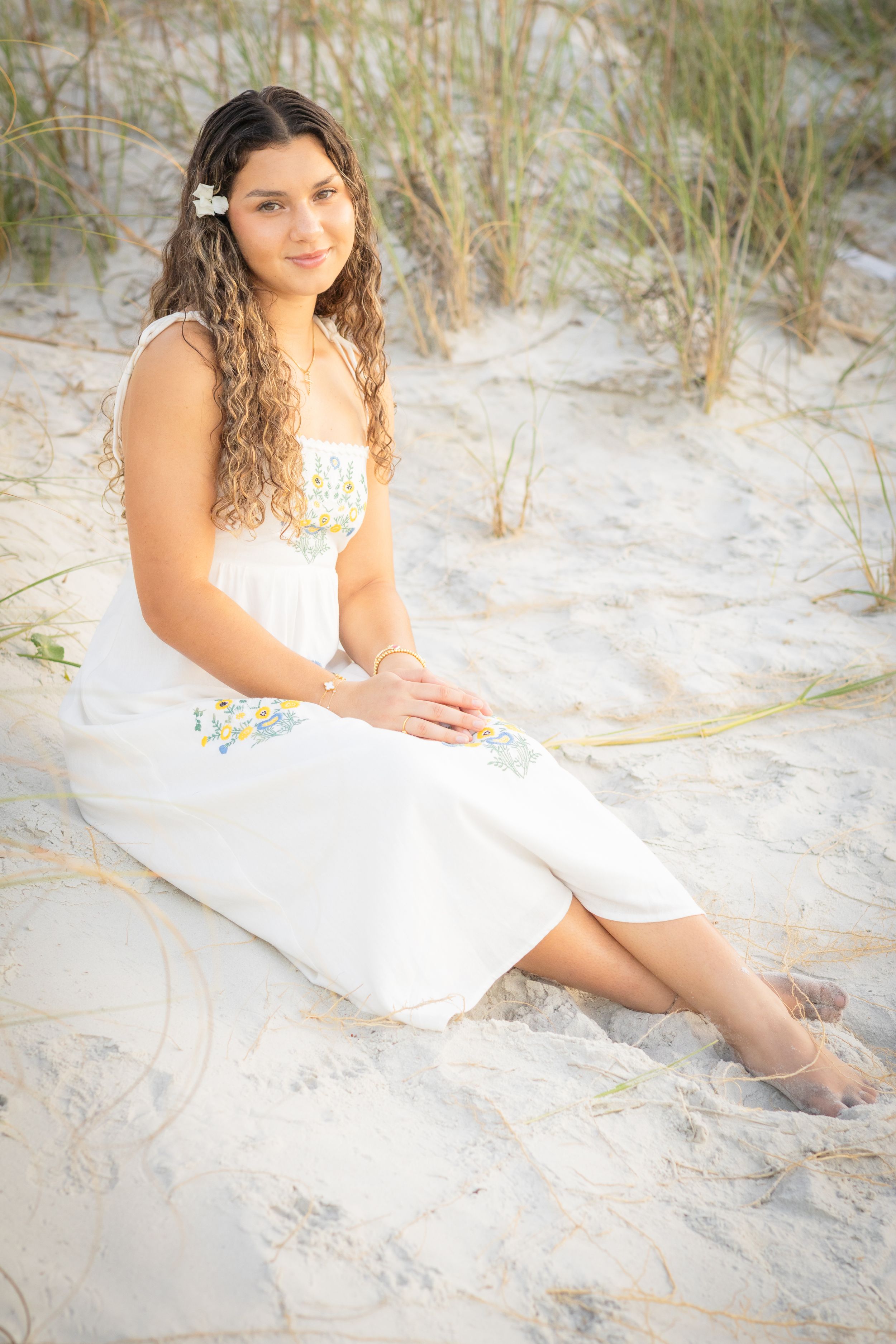 Young woman in white embroidered dress sitting on sandy beach dunes with flower in curly hair.