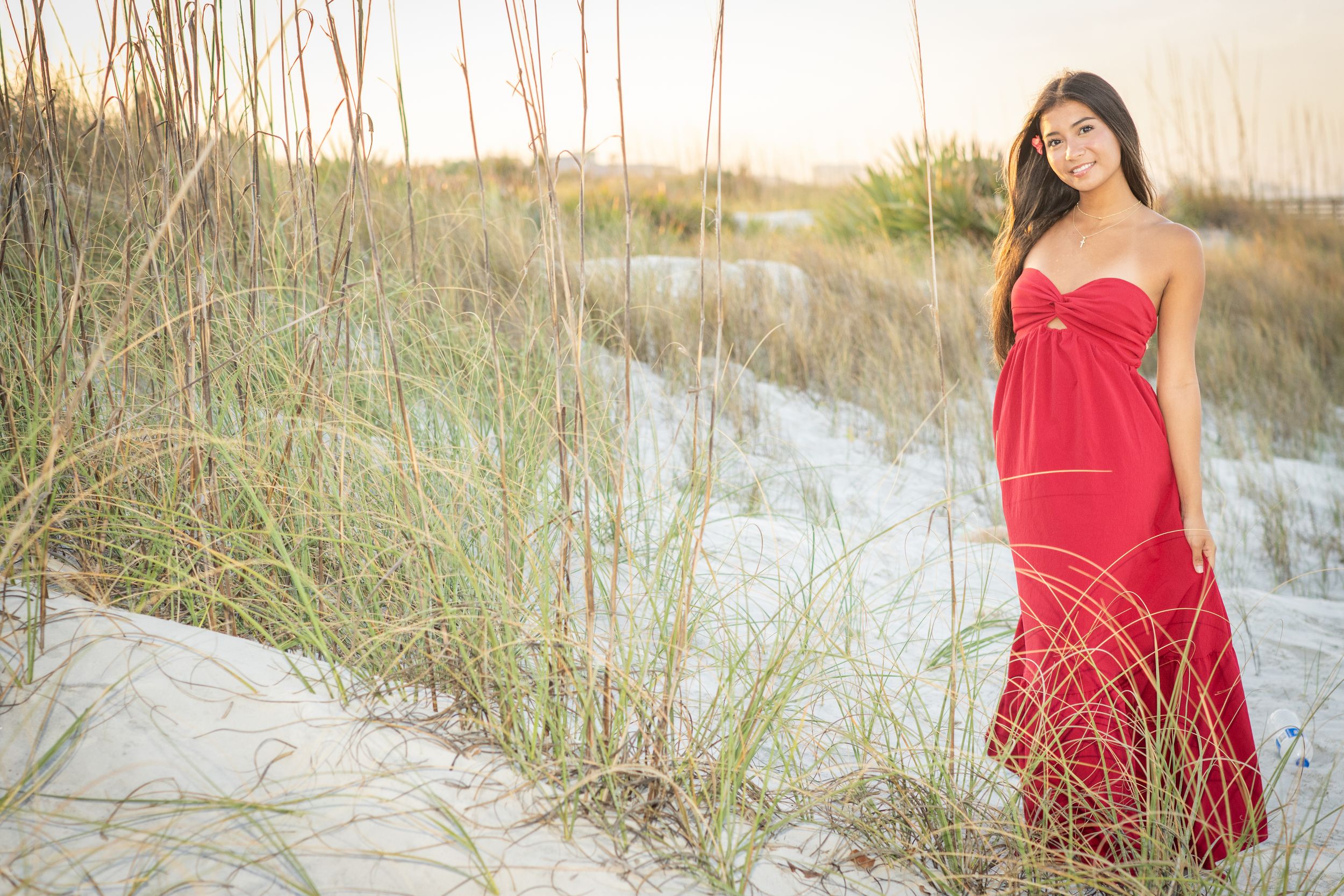 A woman in a red strapless gown poses gracefully among sand dunes and sea grass at sunset on a beach.