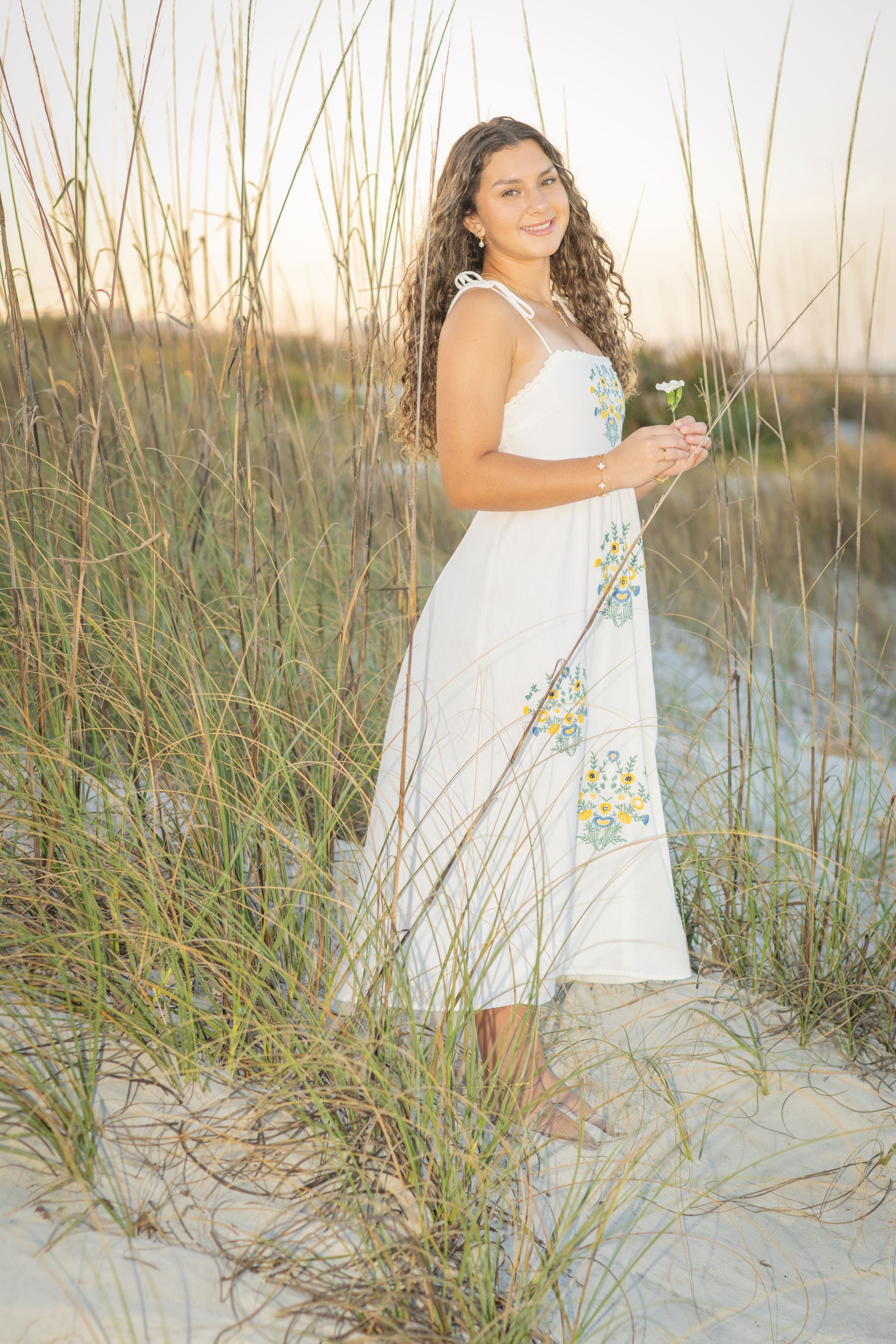 Young woman in white floral sundress standing among beach grass on sandy dunes at sunset, holding a small flower.