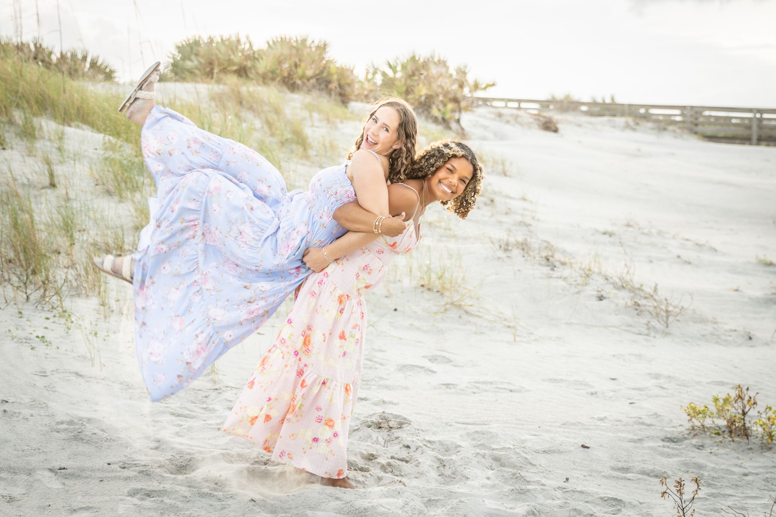 Two women in flowy pastel gowns laugh and play piggyback on a sandy beach dune.