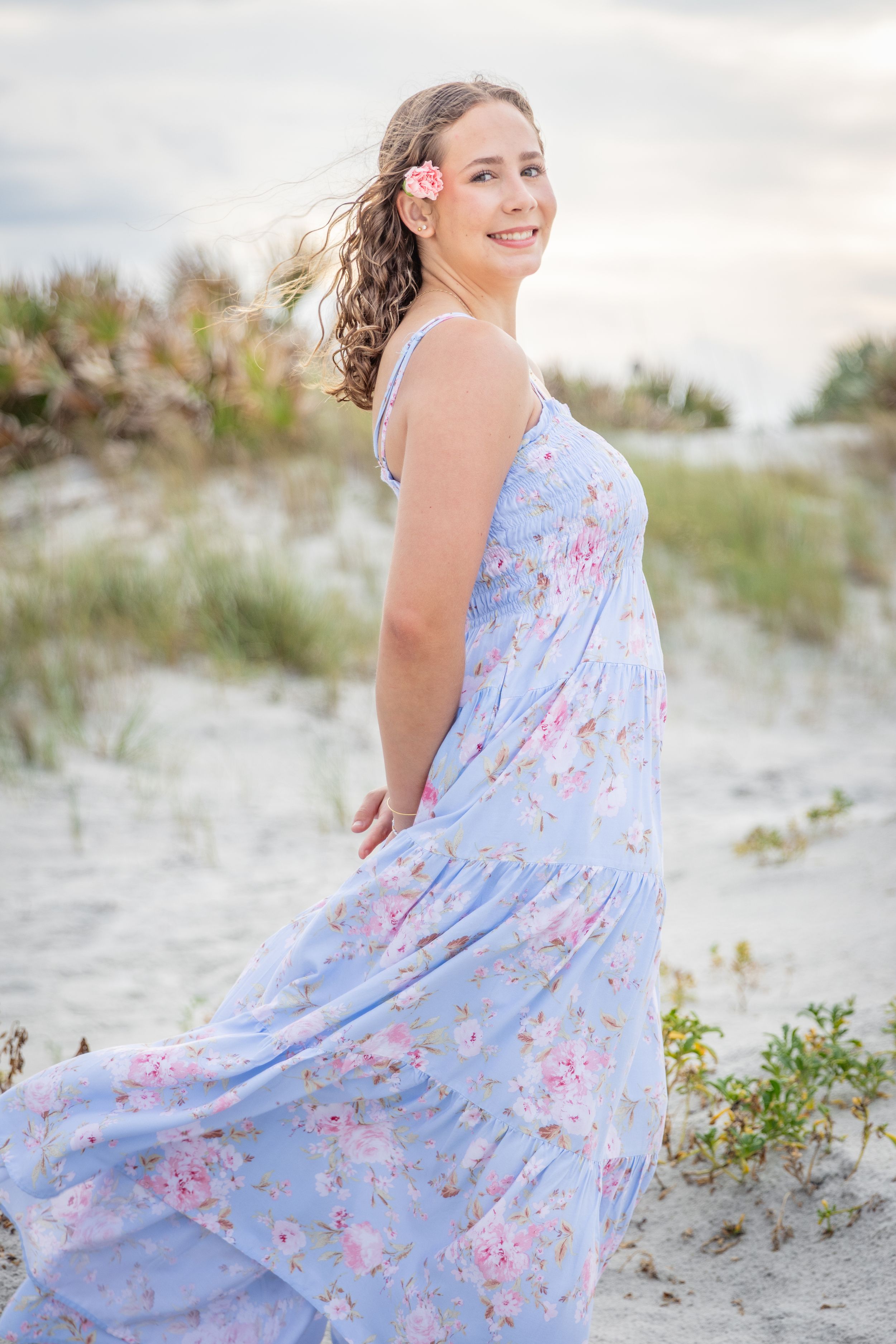 Woman in blue floral maxi dress with pink flower in hair smiling on sandy beach dunes at sunset.
