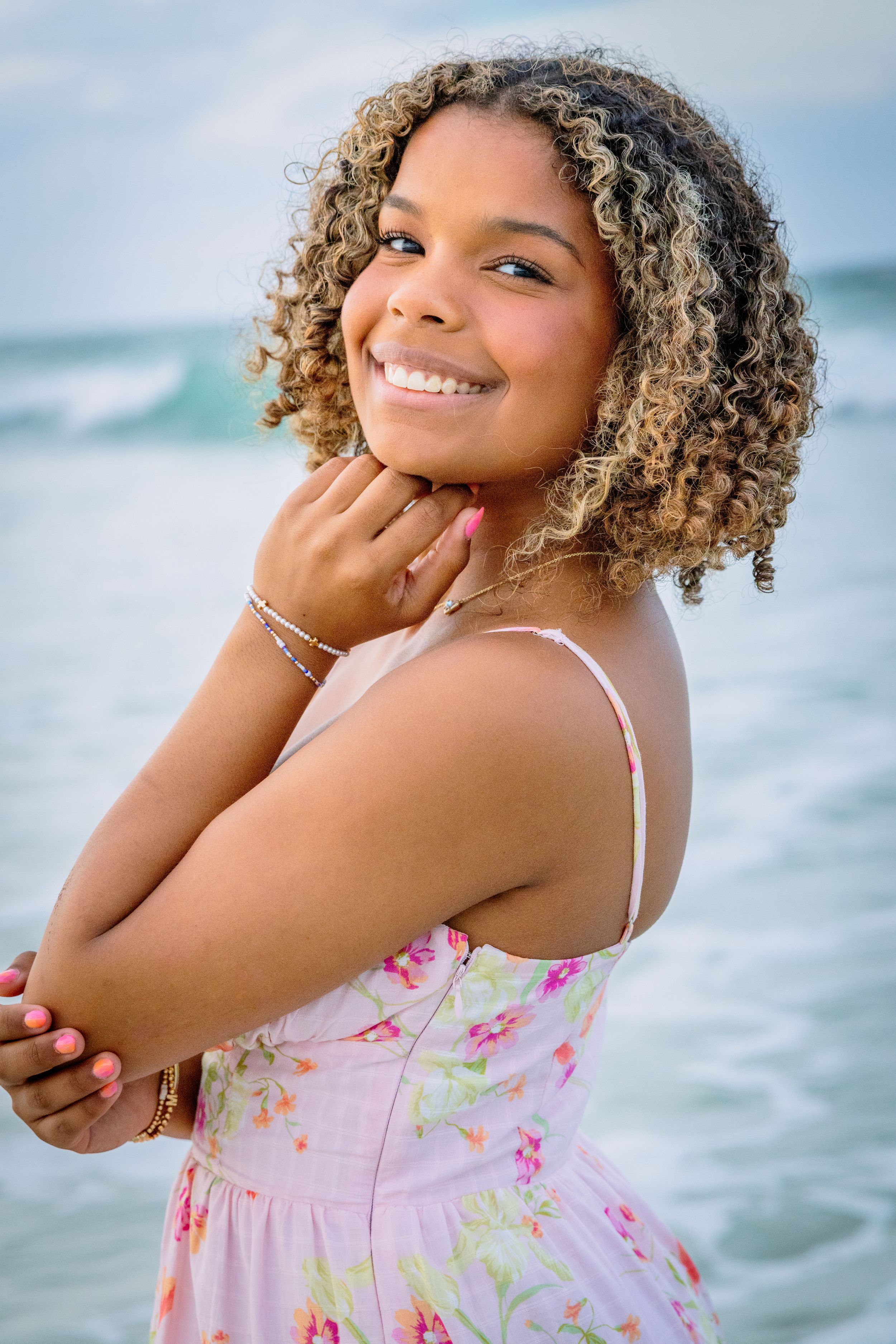 Smiling young woman with curly blonde highlighted hair in a floral pink dress posing at the beach.