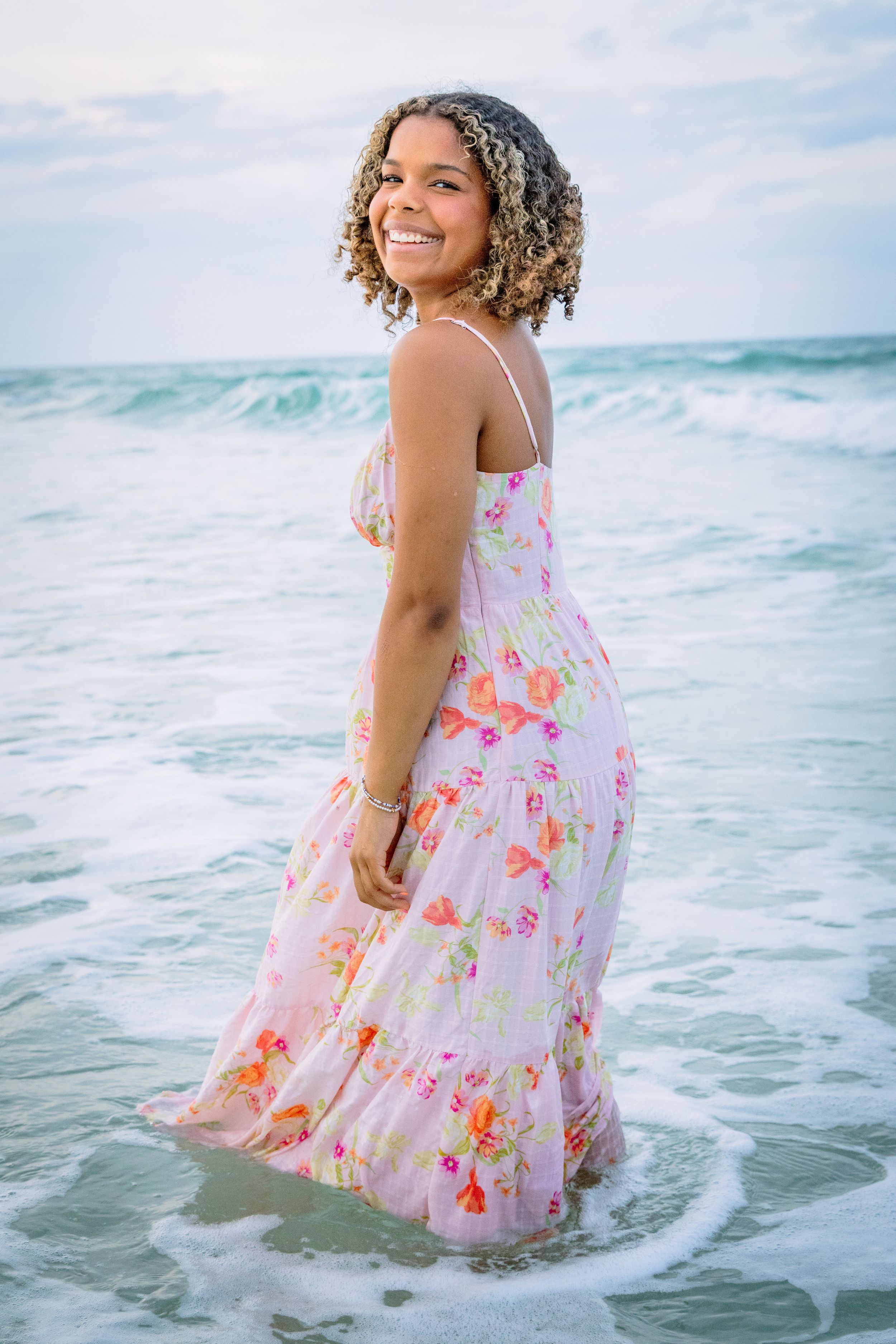 Smiling young woman in floral maxi dress standing in ocean waves on a beach.