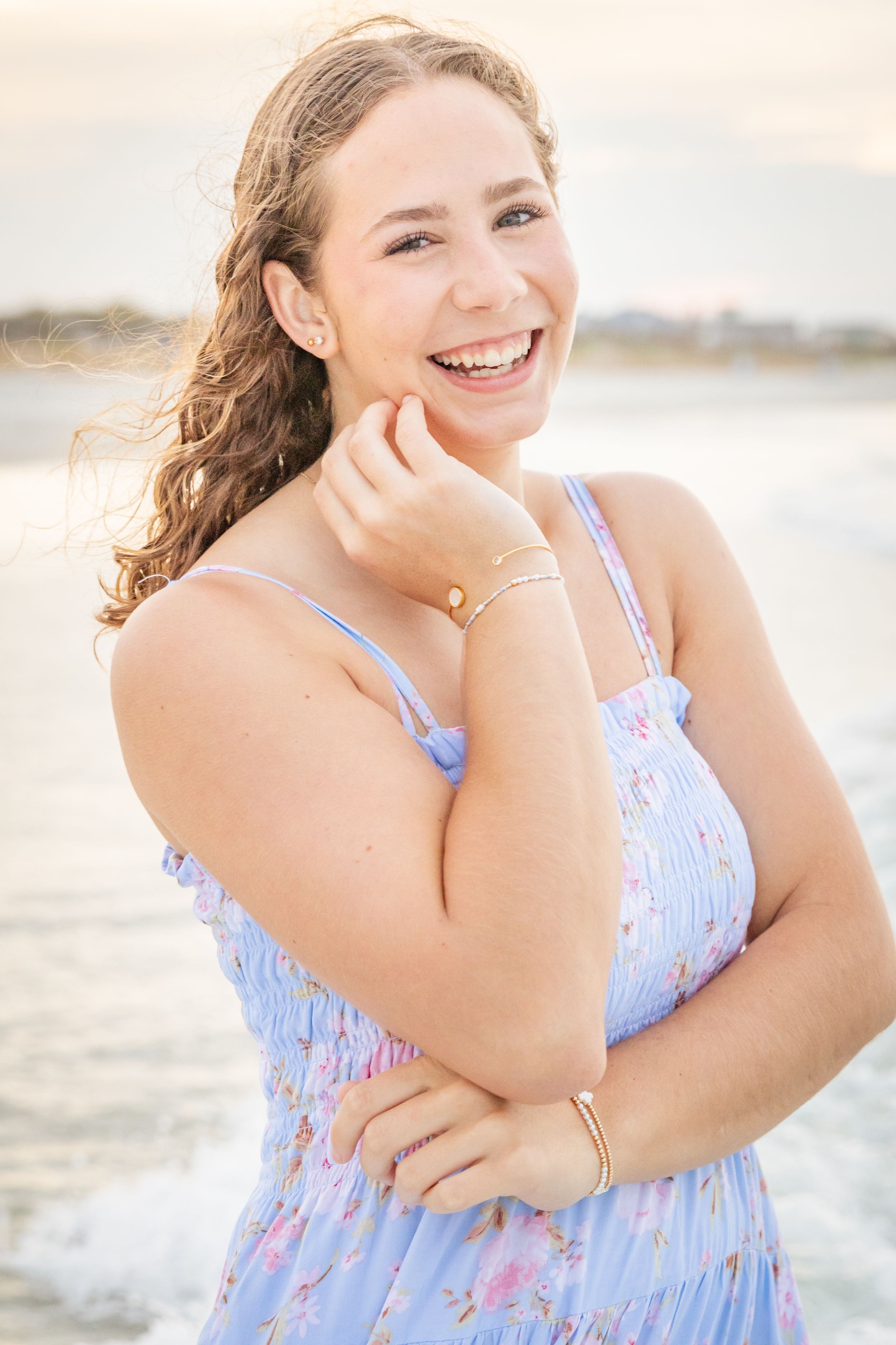 Young woman in a floral blue sundress smiling at the beach during golden hour with ocean waves behind her.