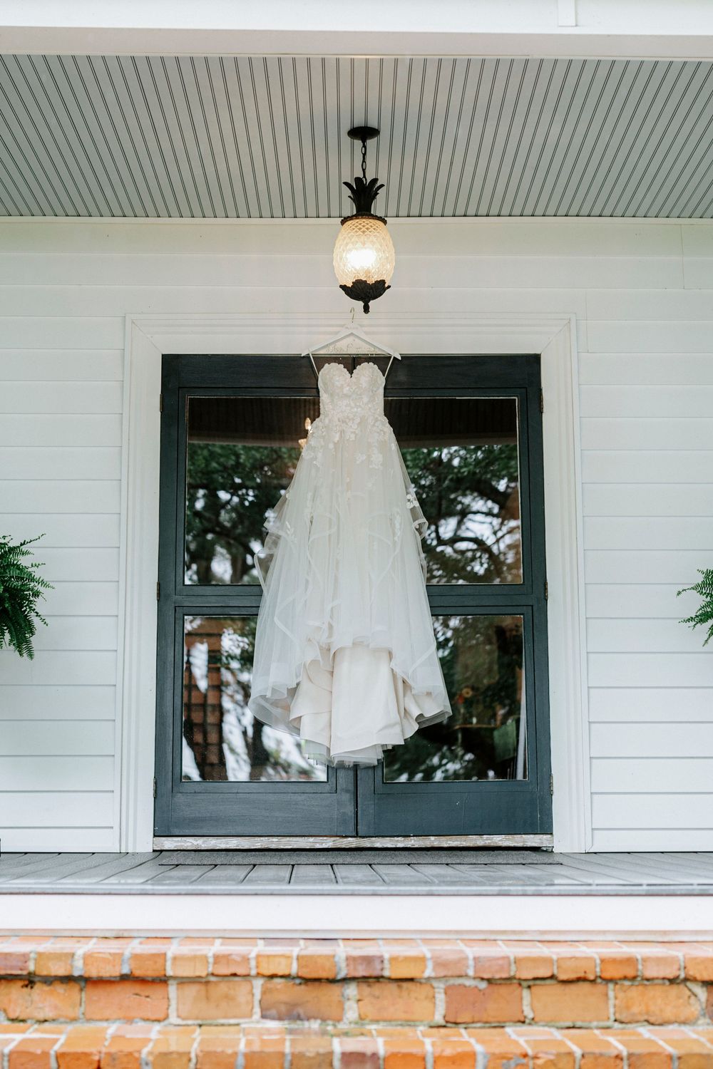 A white strapless wedding gown hanging from a pineapple light fixture on a charming Southern home's front porch.