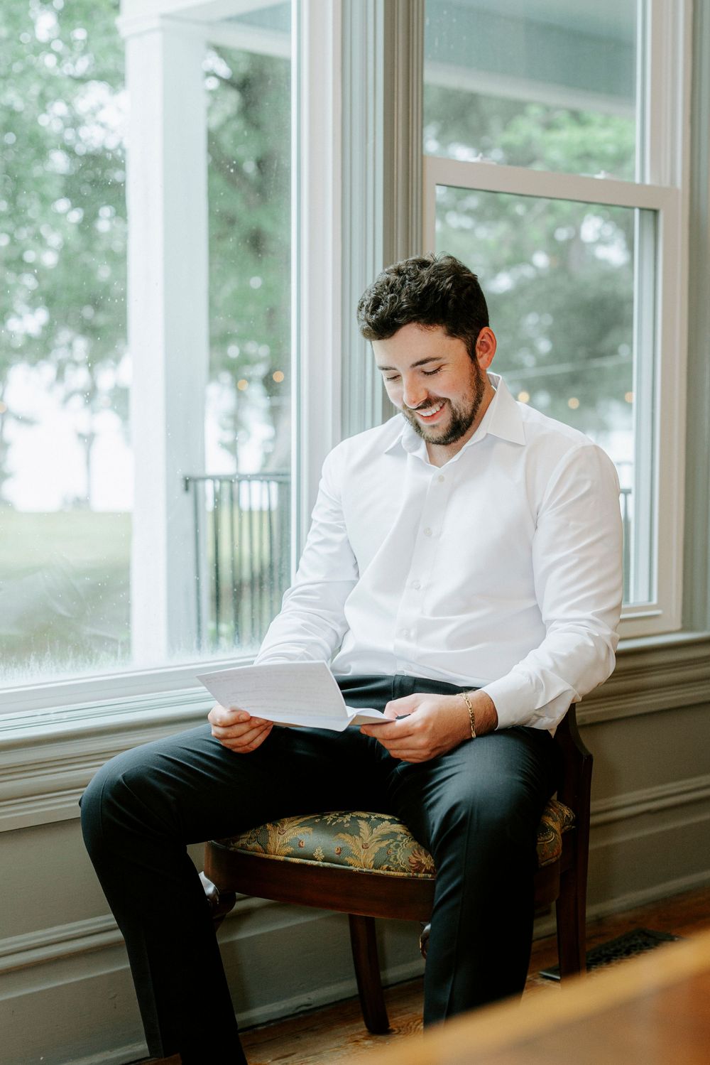 Smiling groom in white dress shirt reads a letter while seated by a bright window on his wedding day.