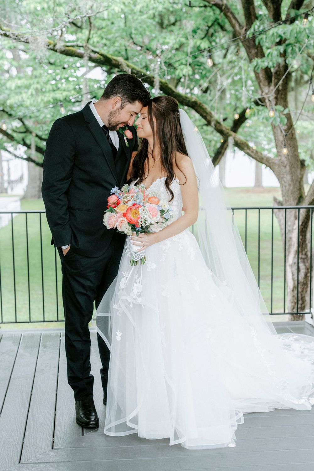 Bride and groom share an intimate moment on a deck, bride holding colorful bouquet in white ballgown with veil.