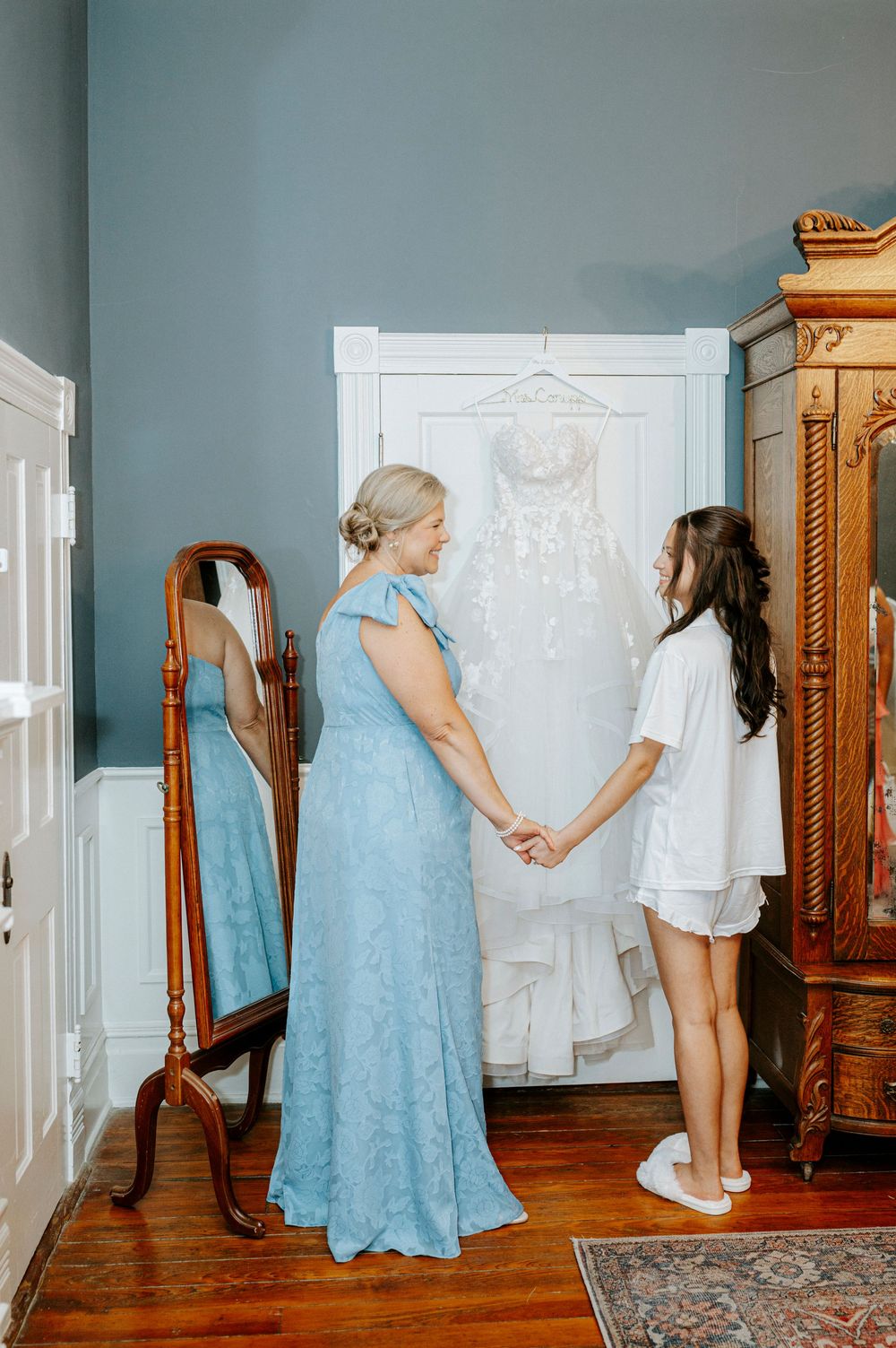 Bride and mother holding hands in blue dress before wedding ceremony with gown displayed on door in elegant room.