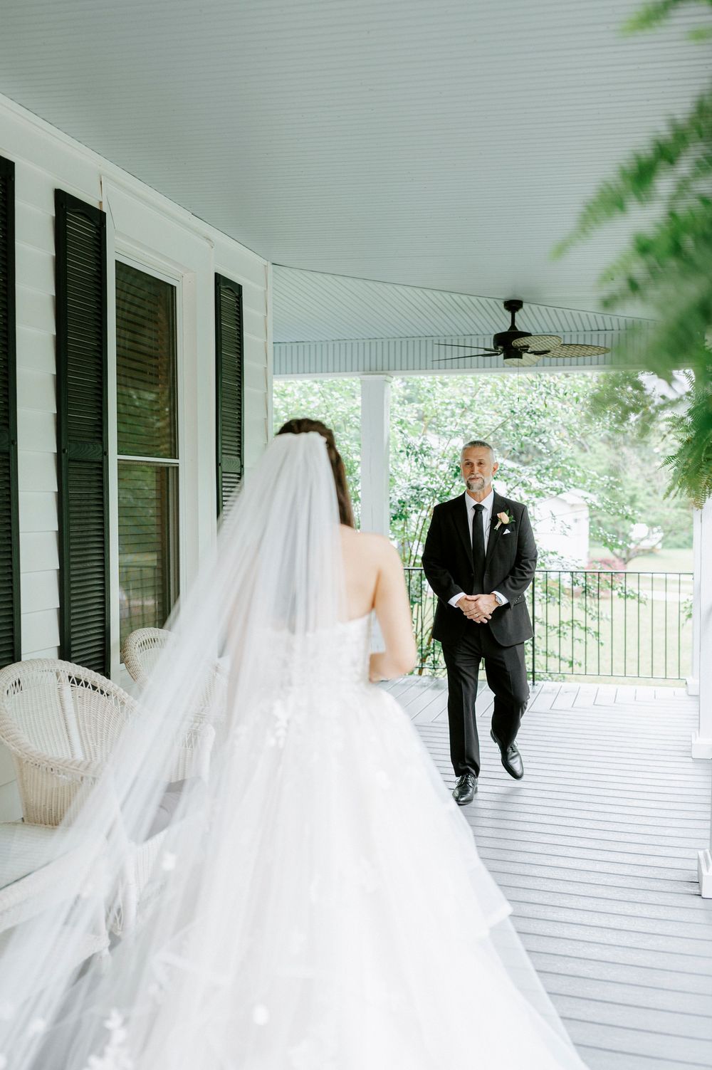 Bride in flowing white gown and veil shares first look moment with groom on classic porch.