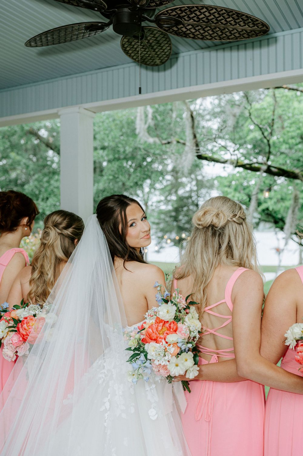 Bride in white gown and veil stands with bridesmaids in pink dresses holding colorful bouquets.