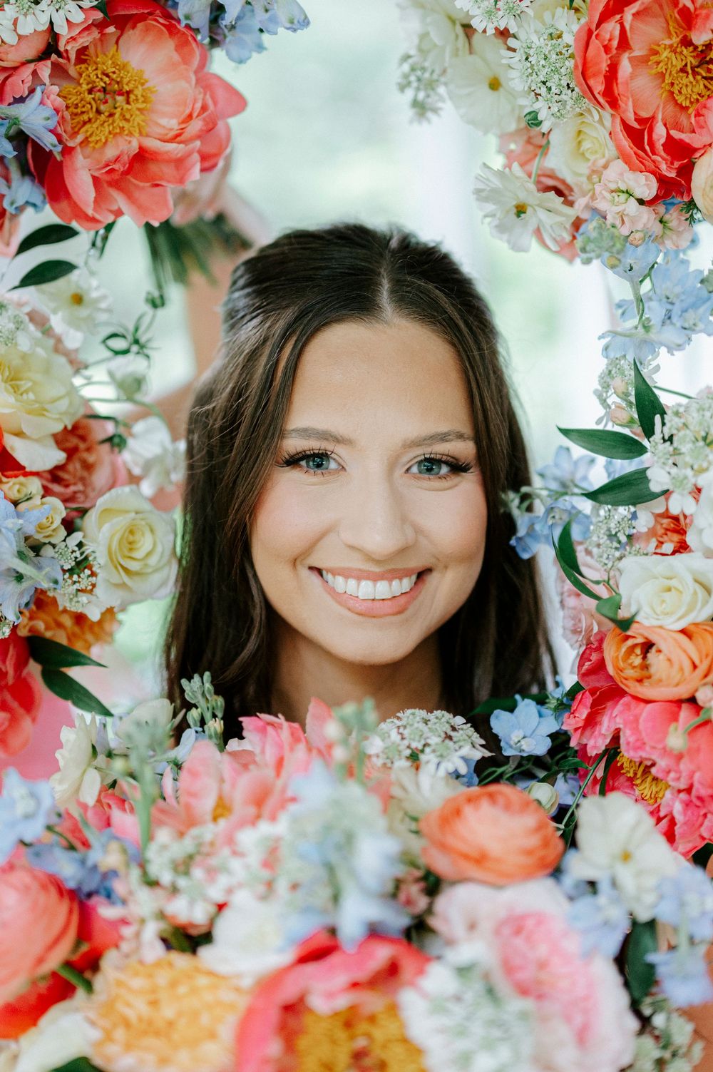 Young woman smiling through a colorful floral frame with pink, coral, and blue flowers at a wedding.