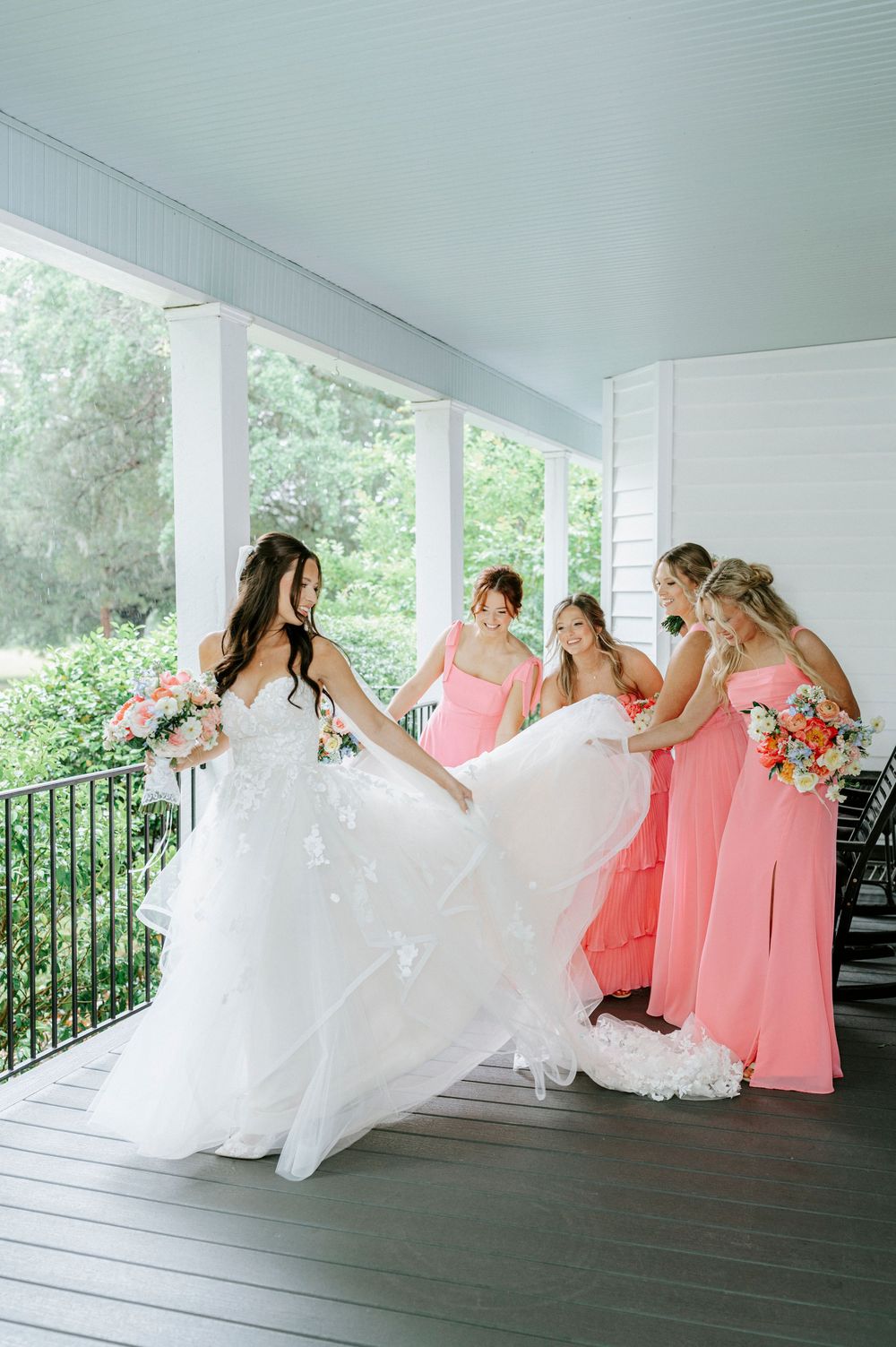 Bride in white lace gown with bridesmaids in coral pink dresses on a porch, adjusting her train.