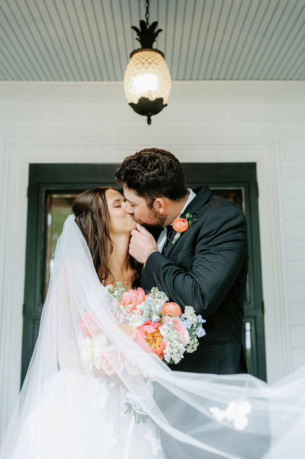 Bride and groom sharing a romantic kiss under a pineapple chandelier, bride holding a colorful bouquet with veil flowing.