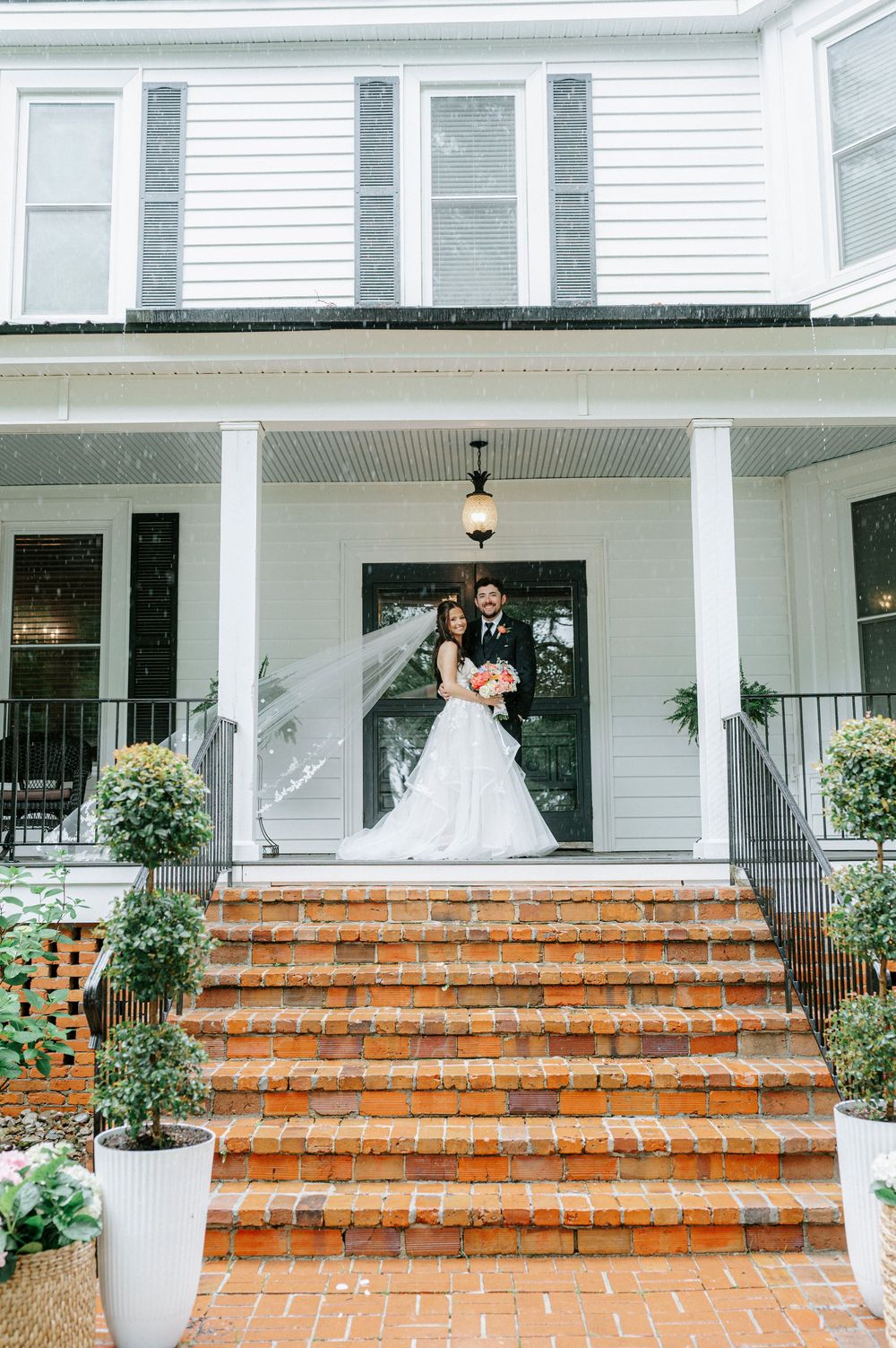Bride and groom pose on brick steps of a white colonial home with columns on their wedding day.
