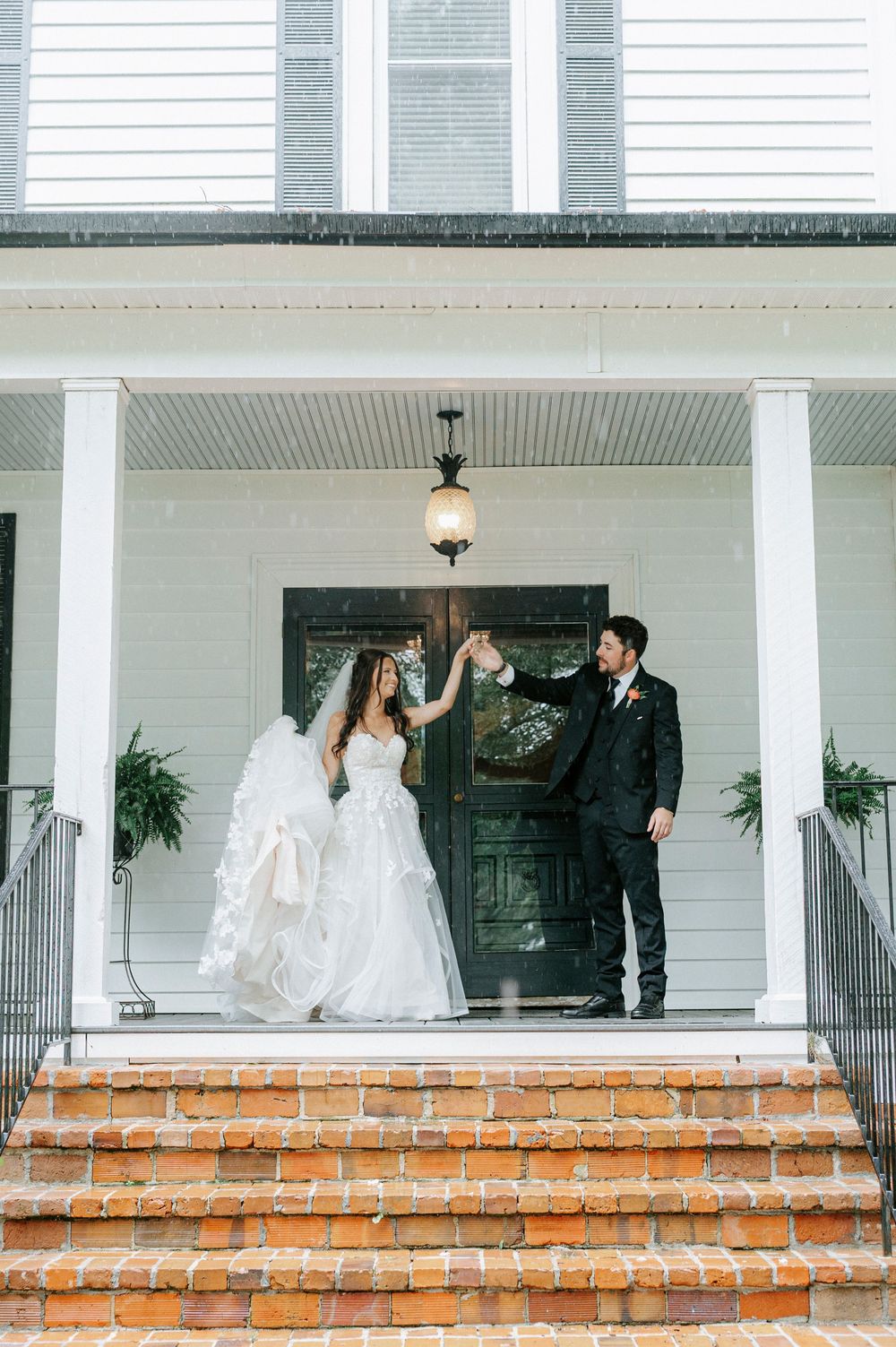 Bride and groom hold hands on brick steps of white Southern home with black door during rainy wedding day.