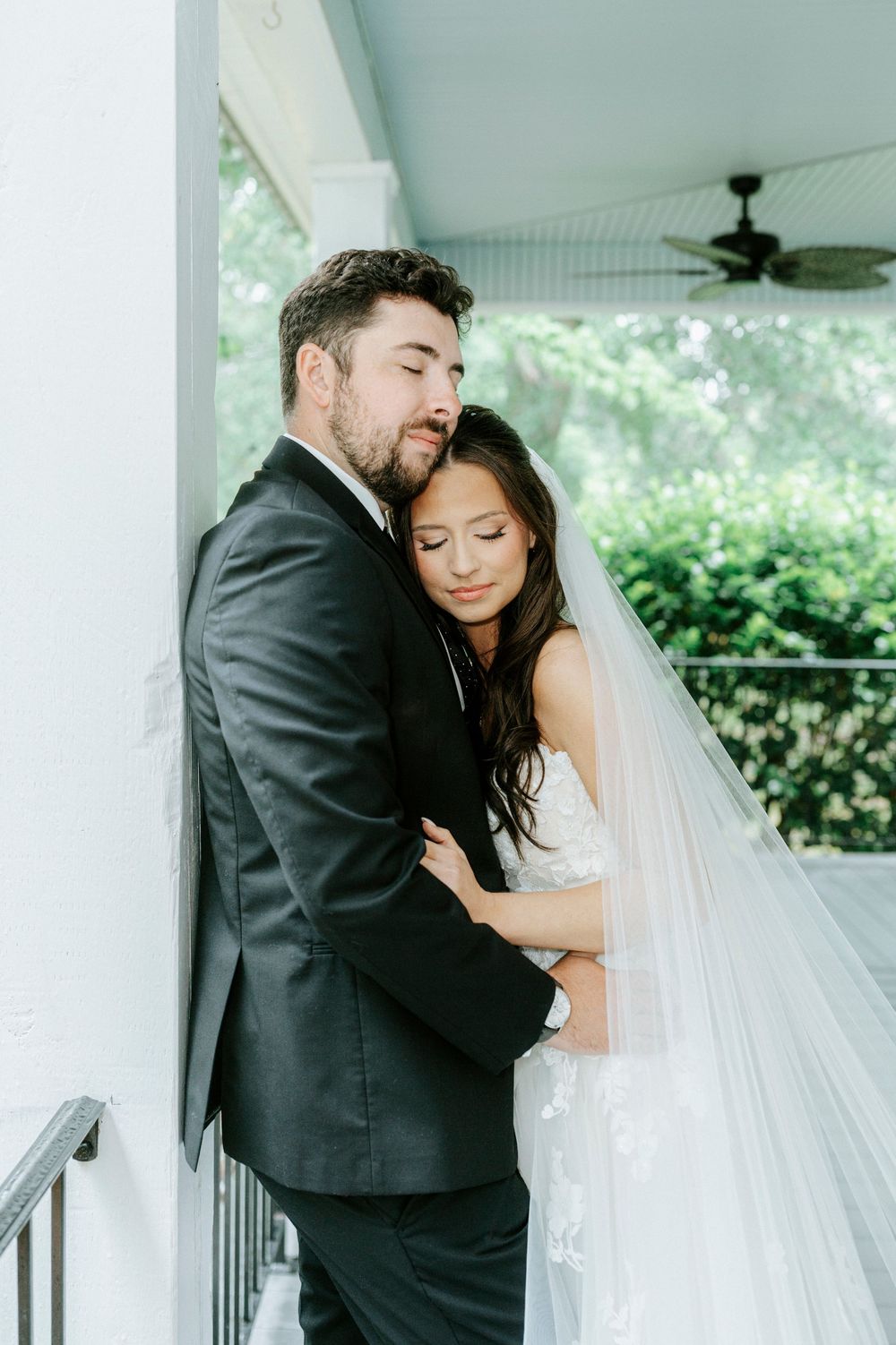 A bride and groom share a tender embrace on a white porch, she in a lace gown with veil, he in a black suit.