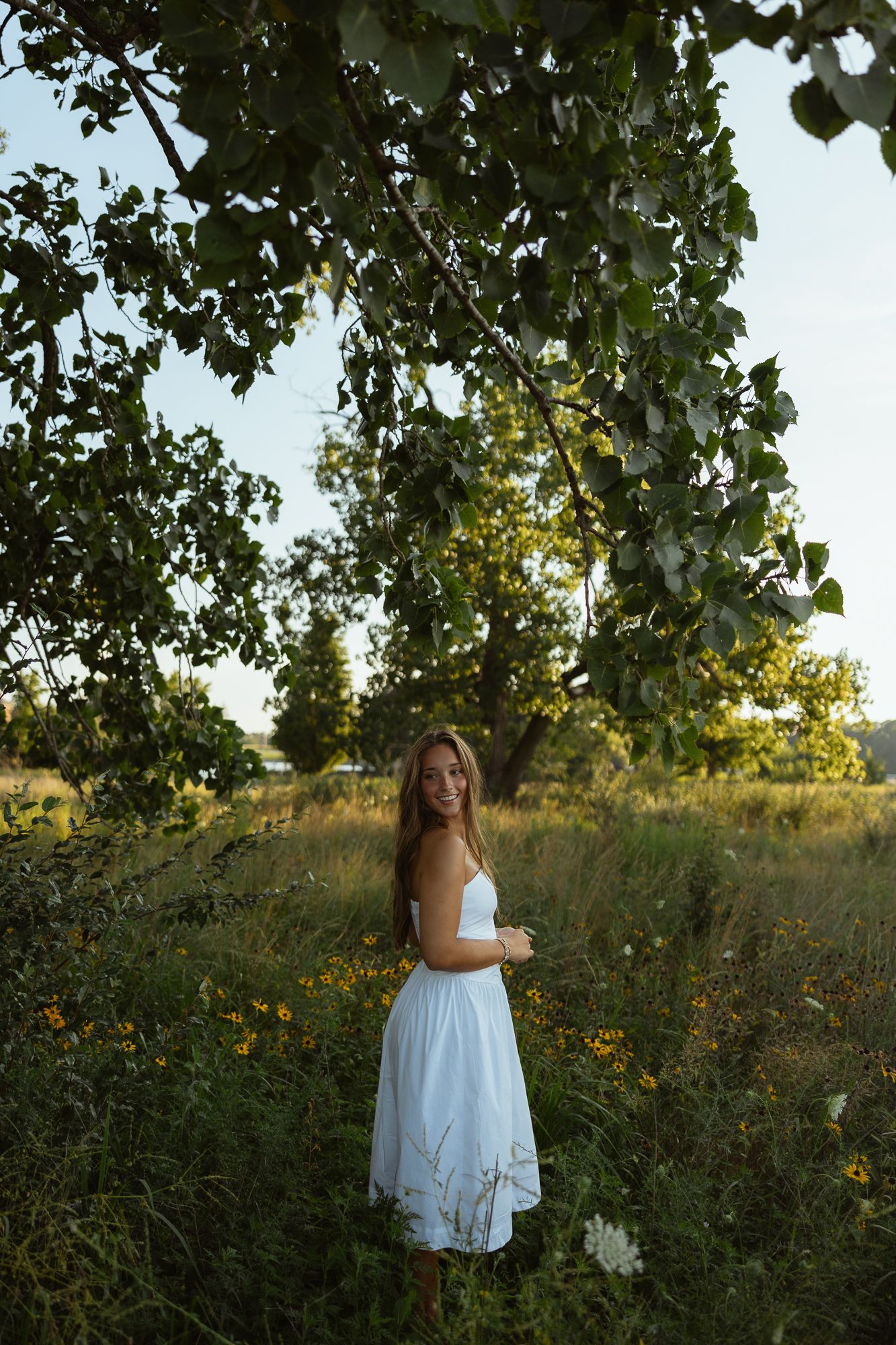Young woman in white sundress standing among wildflowers under tree canopy in golden hour sunlight.
