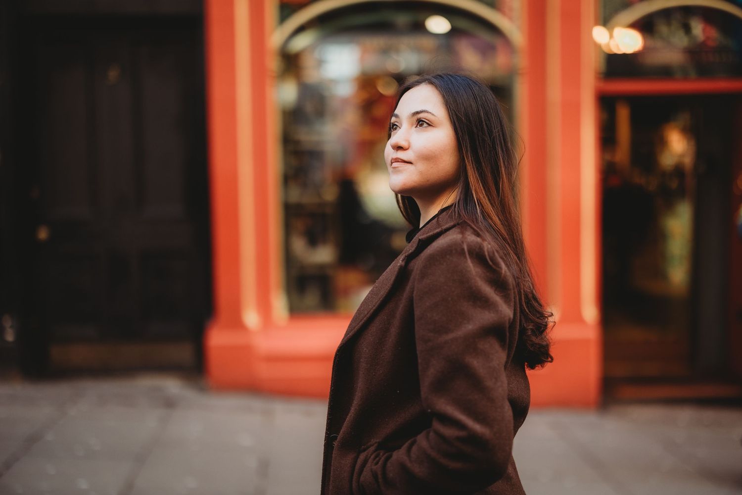 Young woman in brown coat leaning against colorful storefront on a sunny urban street.