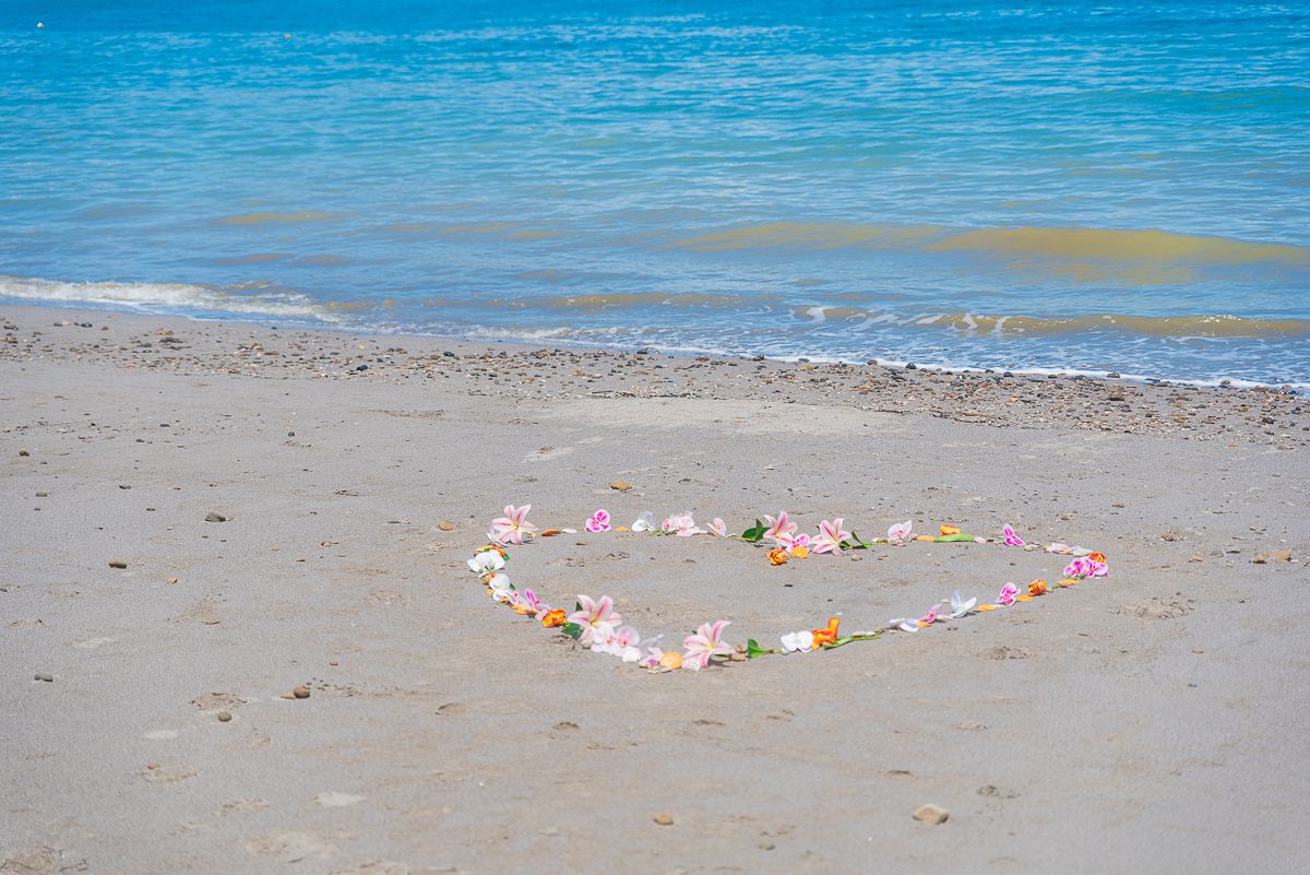 Heart shape made of colorful flowers on sandy tropical beach with turquoise ocean waves in background.