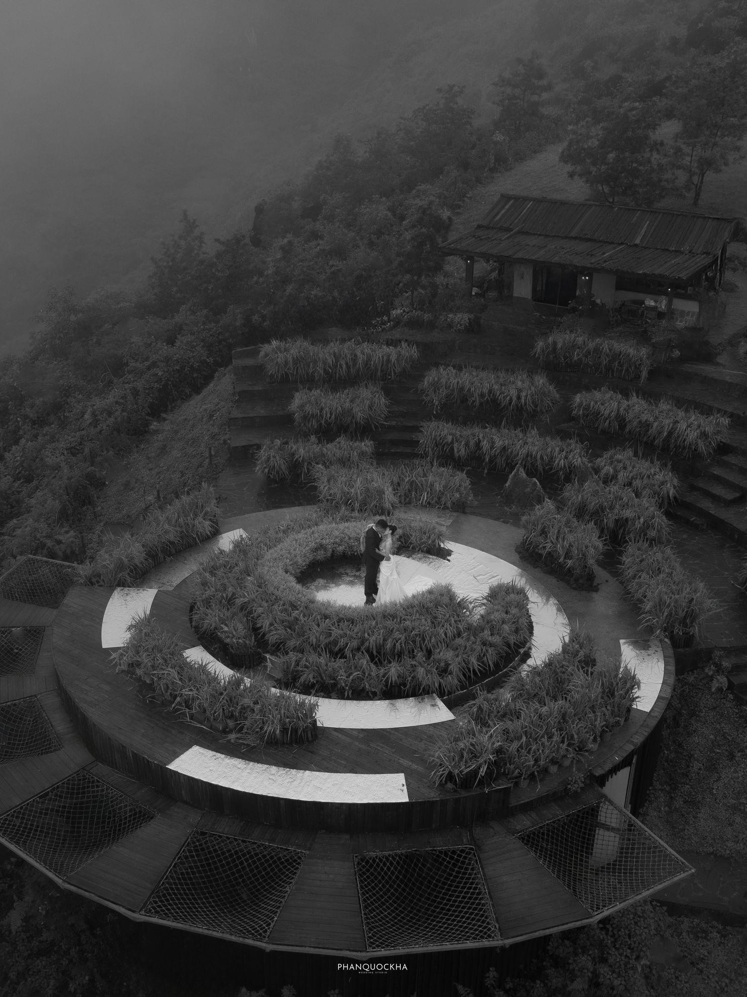 Aerial black and white photo of couple embracing on circular garden structure in misty mountains.