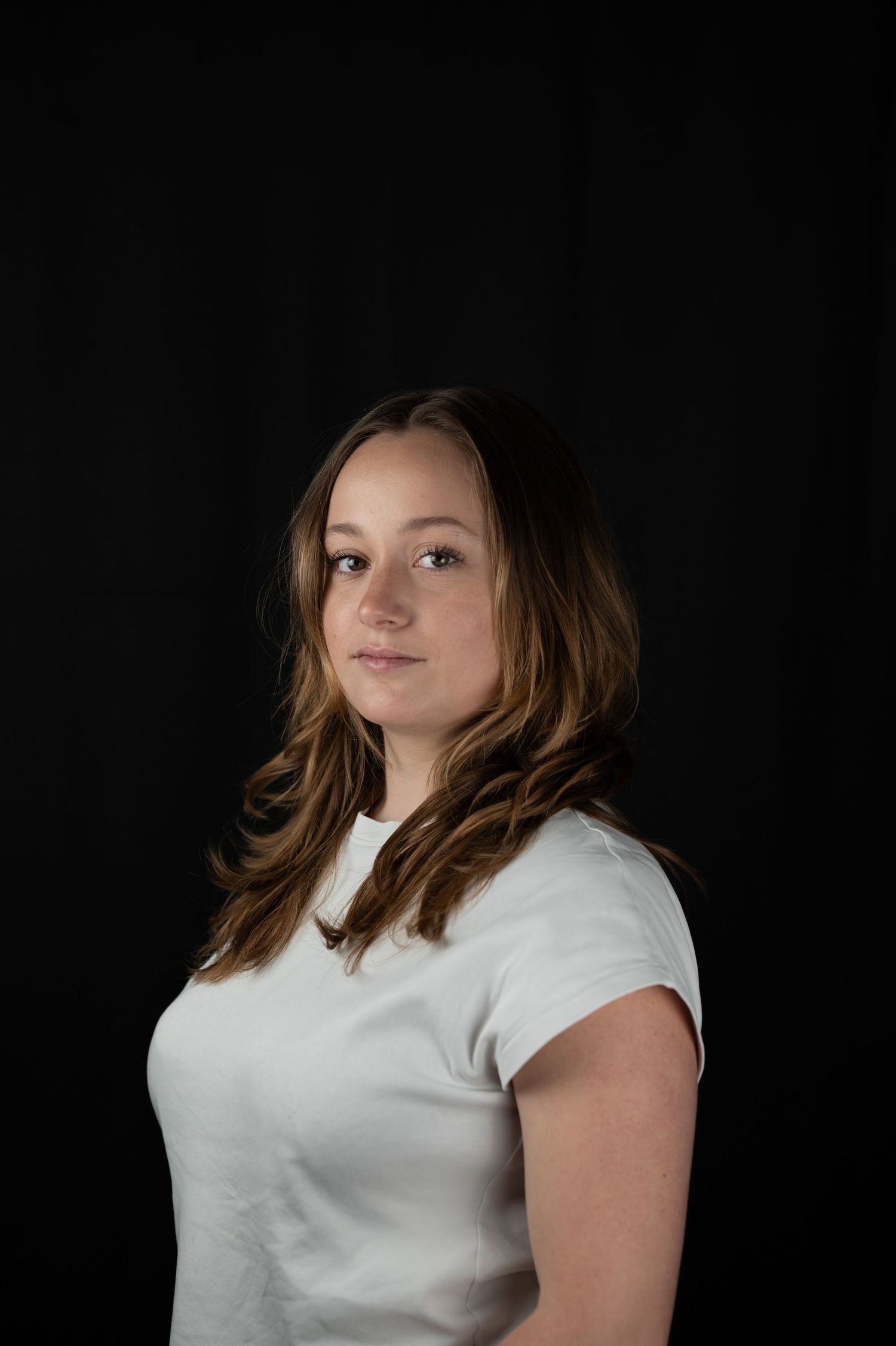 Young woman with long brown hair wearing a white t-shirt posed against a dark black studio background.