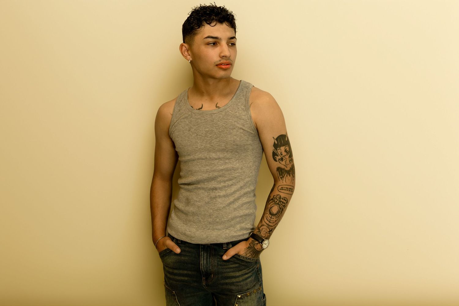 Young man with tattoos wearing a gray tank top and jeans, posing against a beige wall.