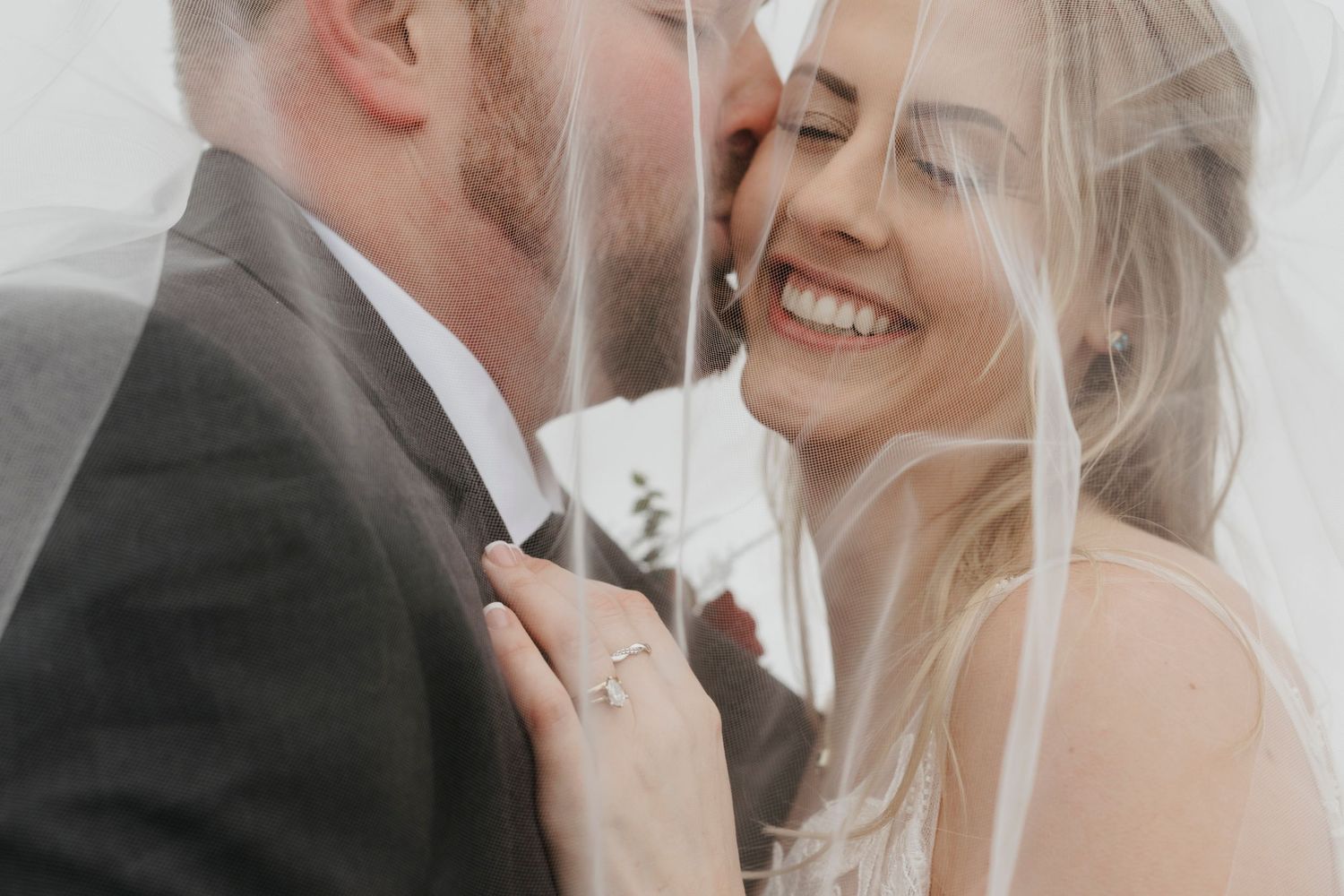A smiling bride and groom share an intimate moment under a wedding veil, with the groom kissing her forehead.