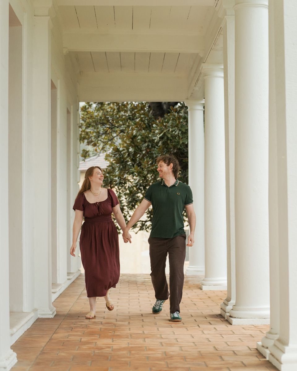 A couple walks hand-in-hand under white columns on a brick path, smiling at each other outdoors in Sydney Milk beach proposal photoshoot