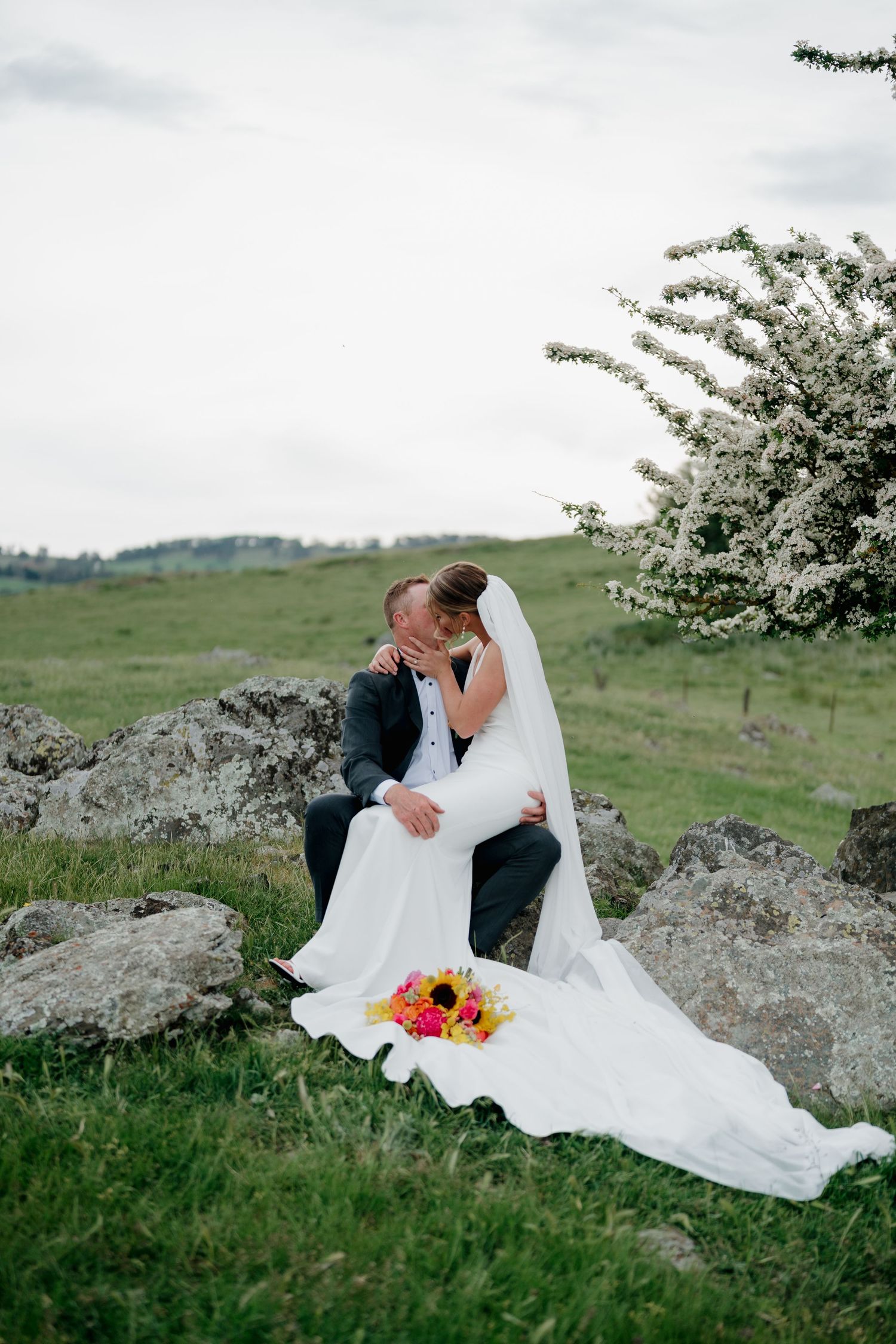 Bride and groom share a romantic moment on rocks in a green field, with a colorful bouquet and blooming tree nearby.