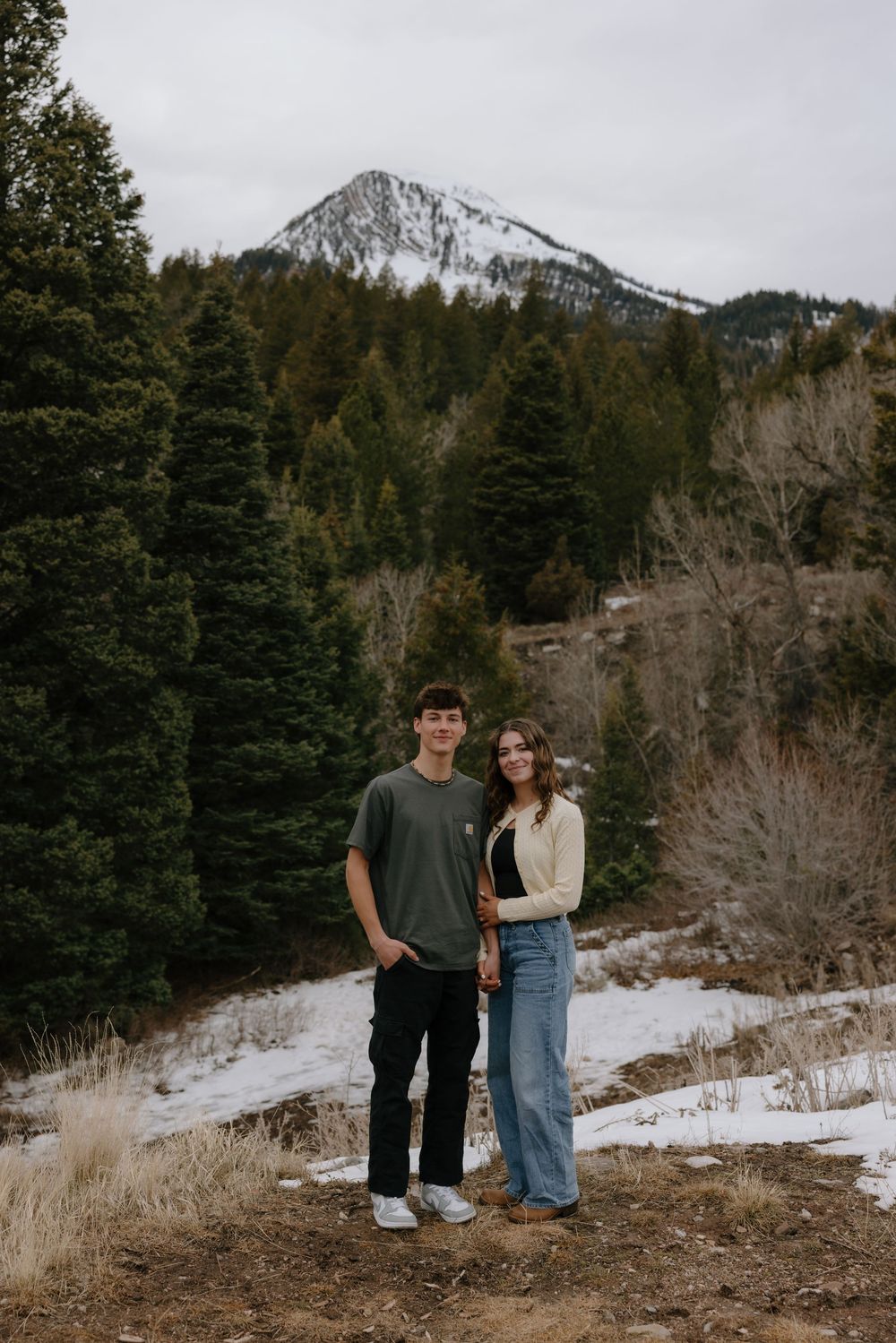 Young couple posing together in snowy mountain landscape with evergreen trees and snow-capped peak behind them.