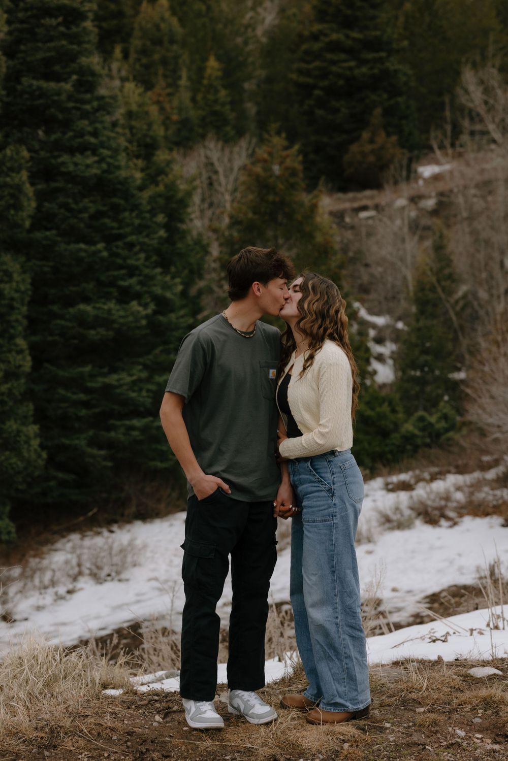 Couple sharing a kiss in a snowy mountain forest setting, man in green tee and woman in cream top and jeans.