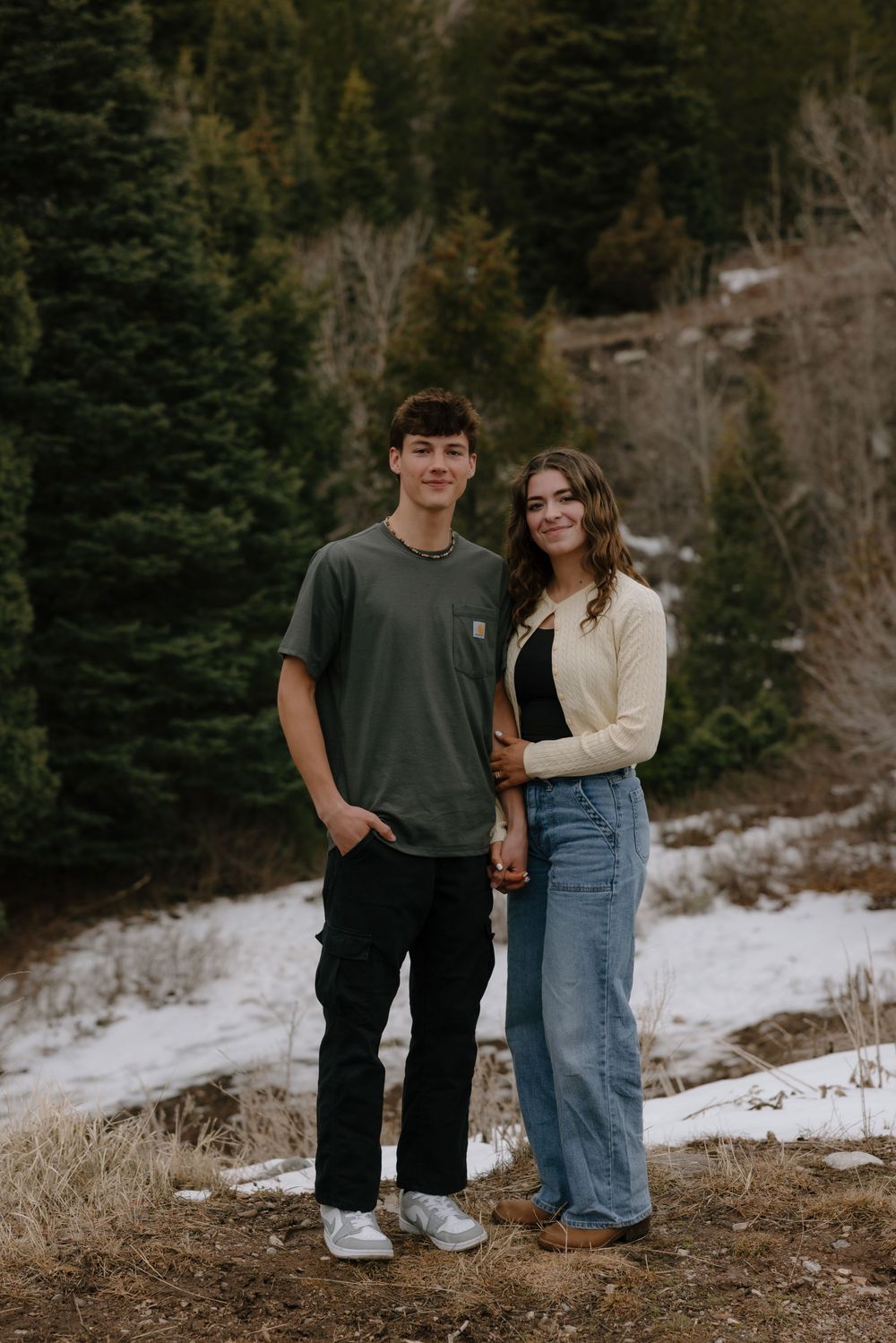 Young couple posing together in a snowy mountain forest setting, wearing casual winter outfits.