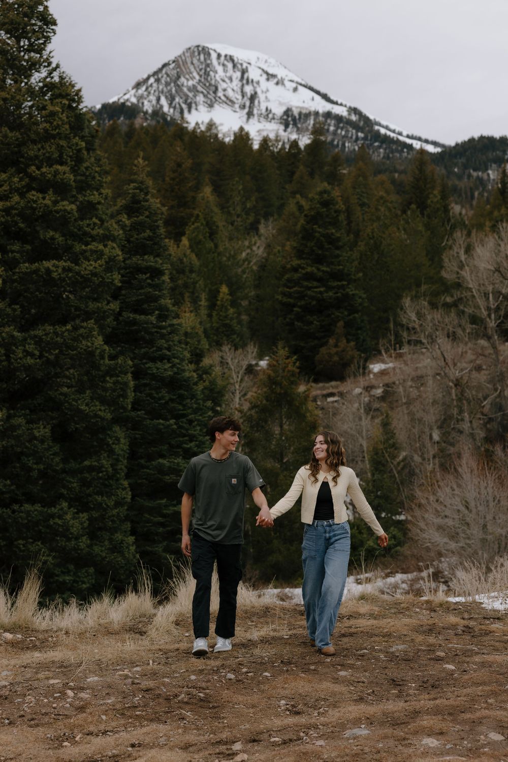 Couple holding hands walking on a mountain trail with snow-capped peaks and evergreen trees in the background.
