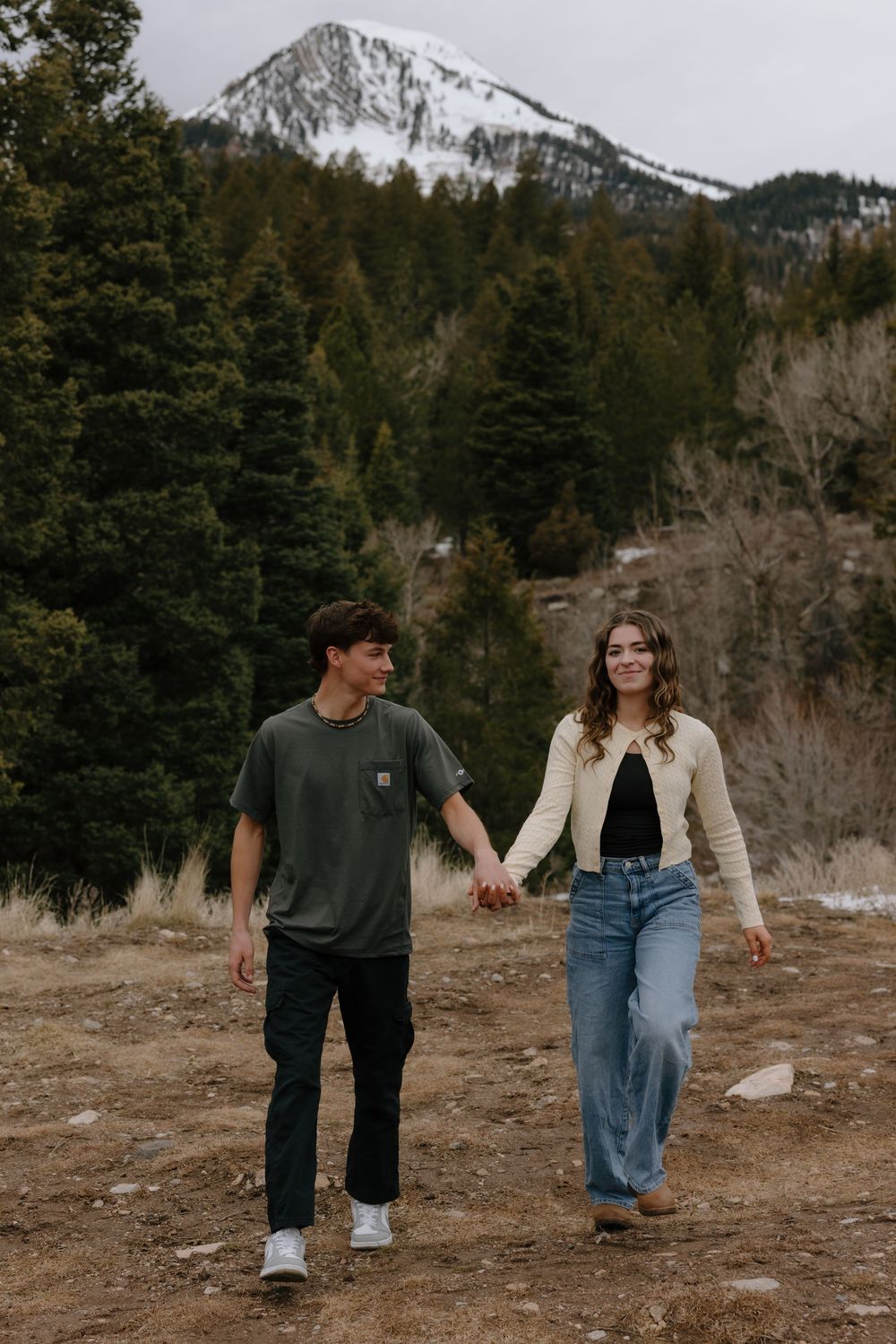 A couple holds hands while walking through a mountain landscape with evergreen trees and a snow-capped peak behind them.