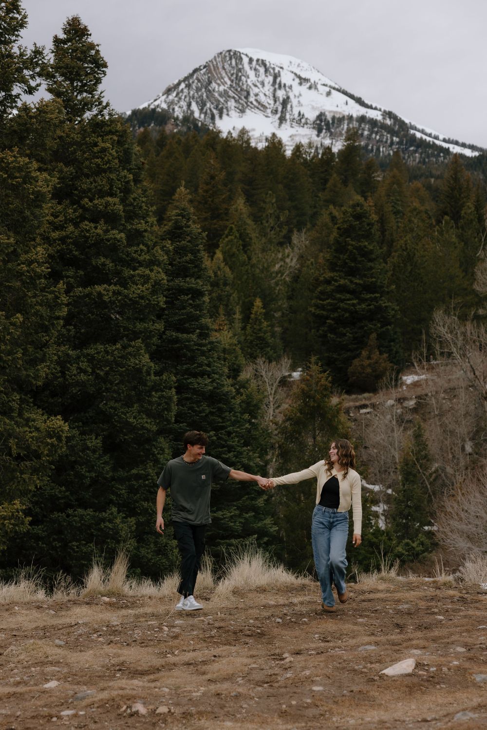 Young couple holding hands walking on a mountain trail with snow-capped peak and evergreen forest backdrop.