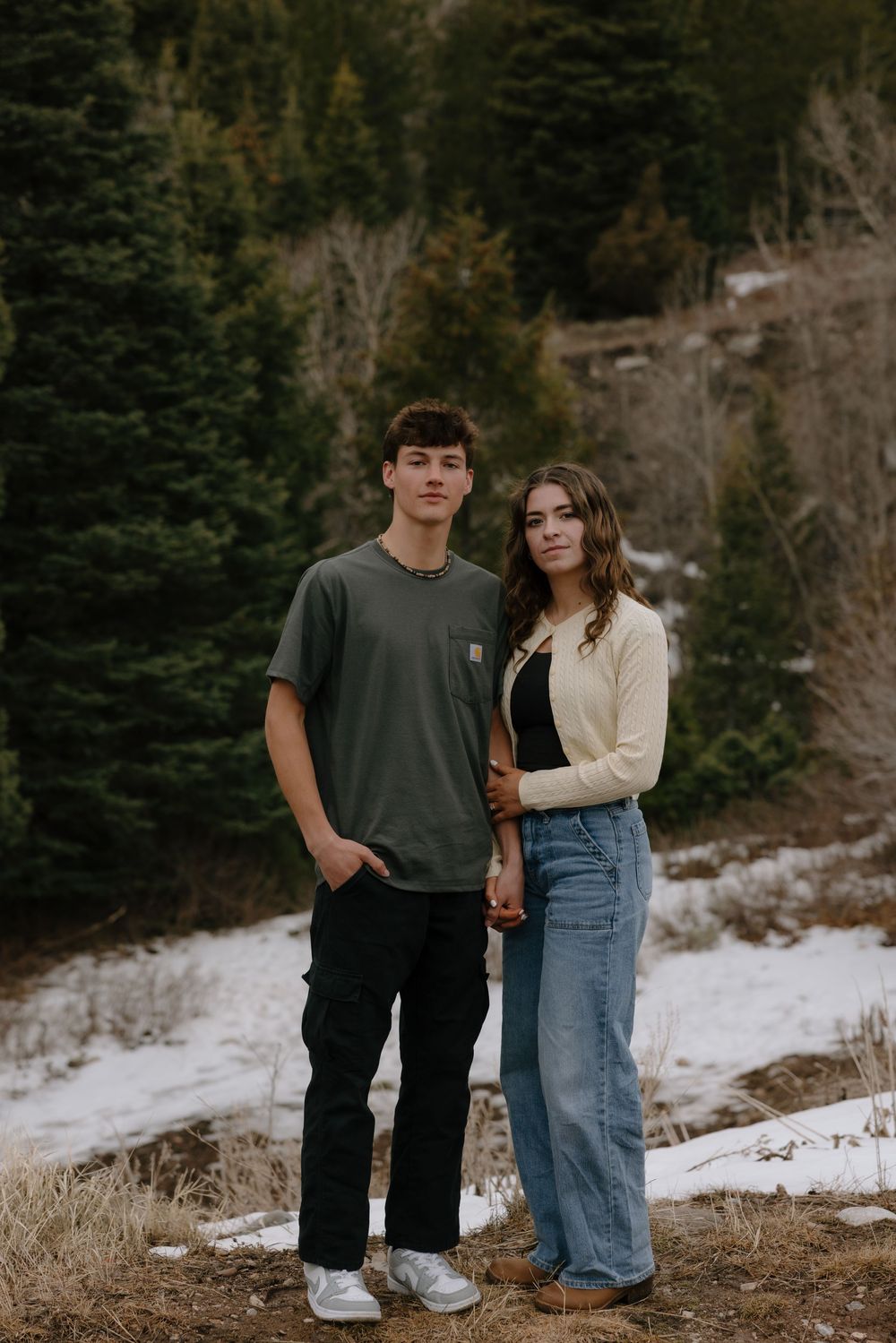 A young couple poses together in a snowy mountain forest setting, wearing casual winter outfits.