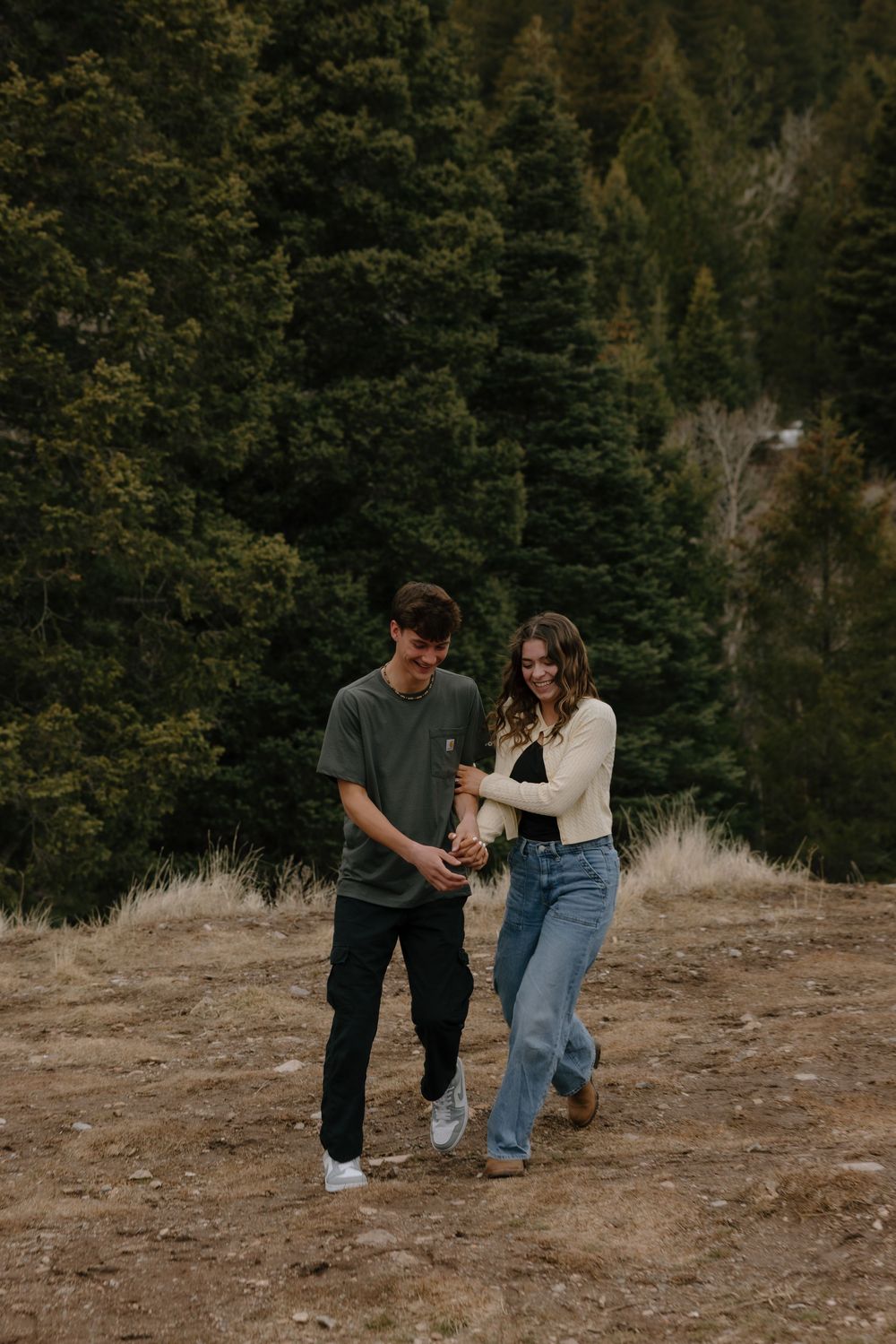 A young couple walks together on a hillside trail surrounded by evergreen trees in a scenic outdoor setting.