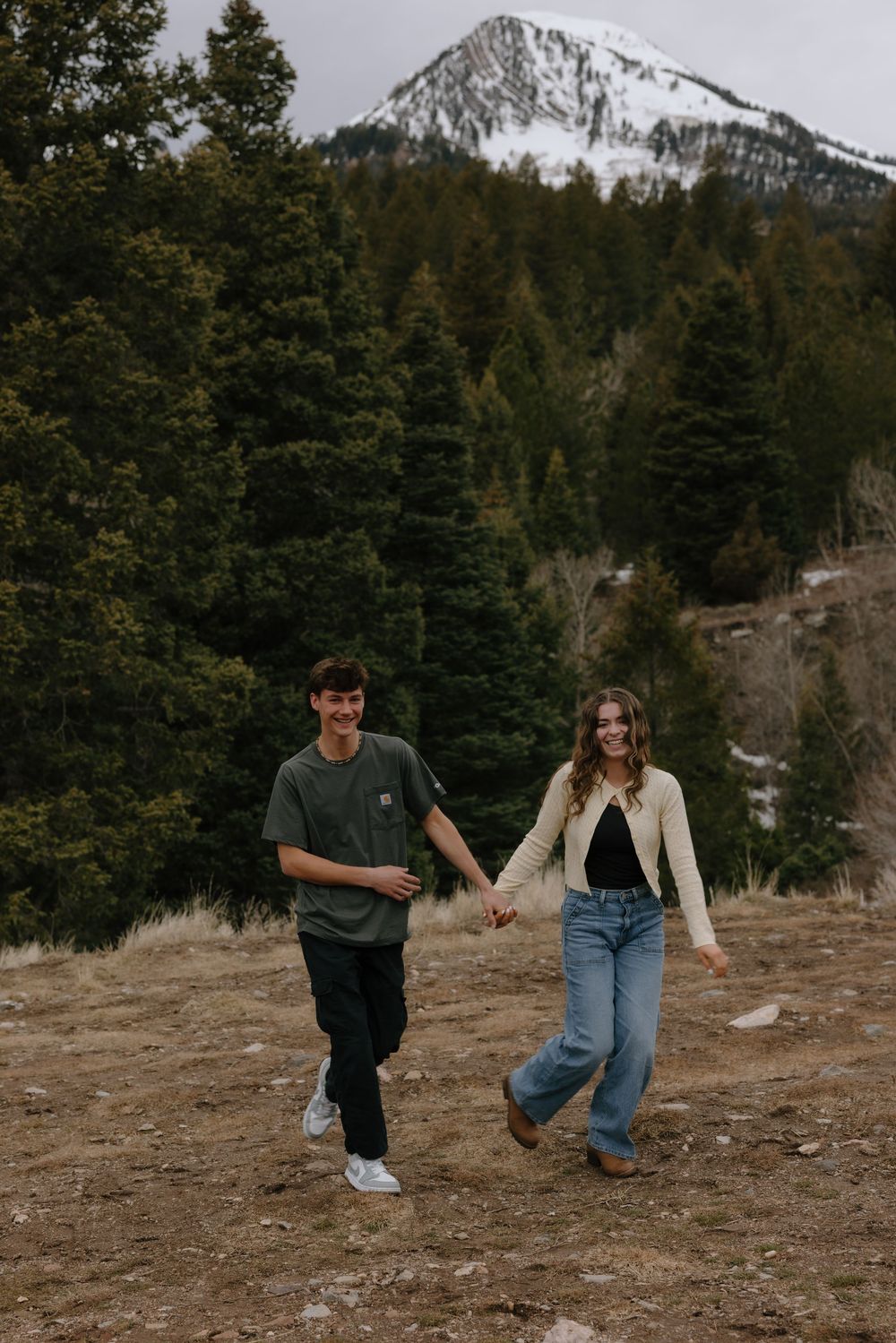 Young couple holding hands walking through a mountain meadow with snow-capped peaks and pine trees in background.
