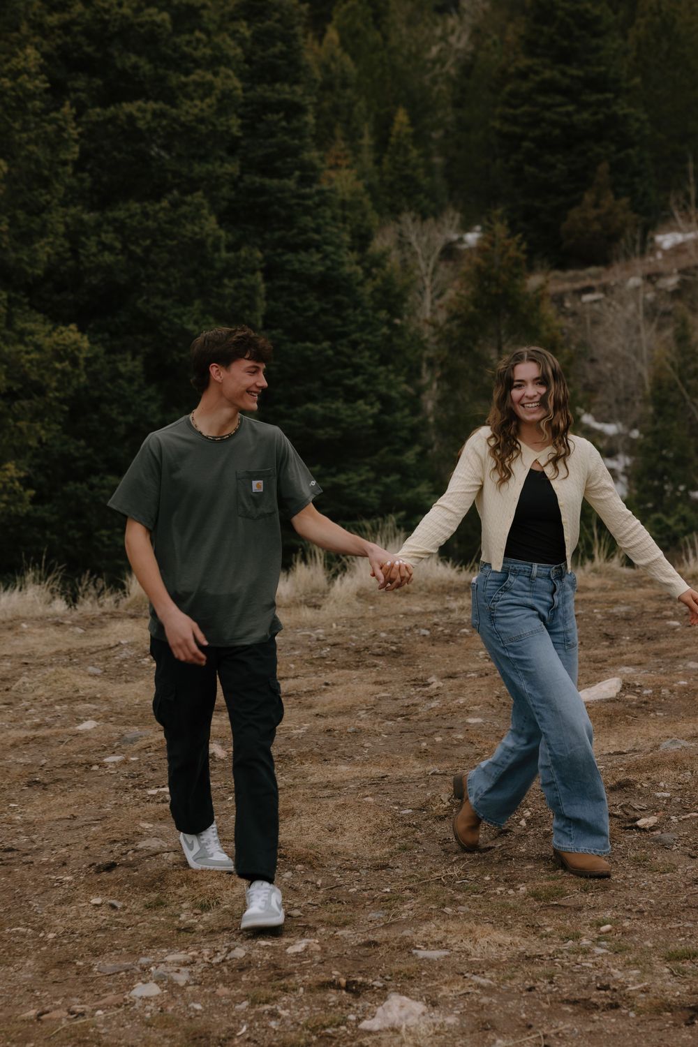 Smiling young couple holding hands and walking together on a hillside with evergreen trees behind them.