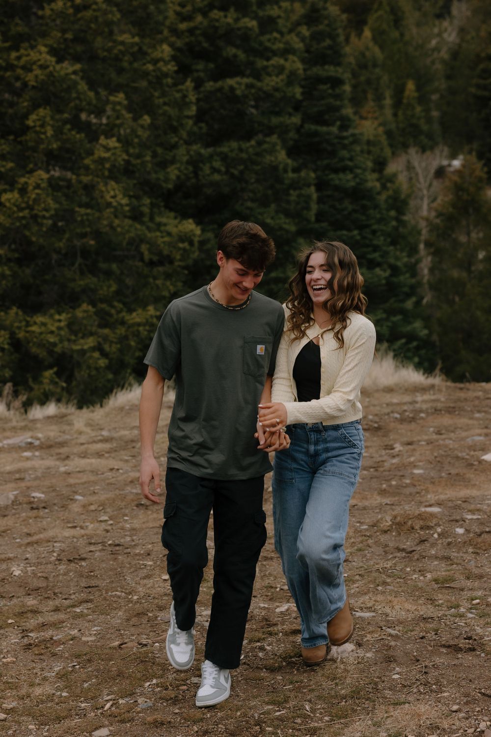 A smiling couple walks hand-in-hand through a scenic outdoor field with evergreen trees in the background.