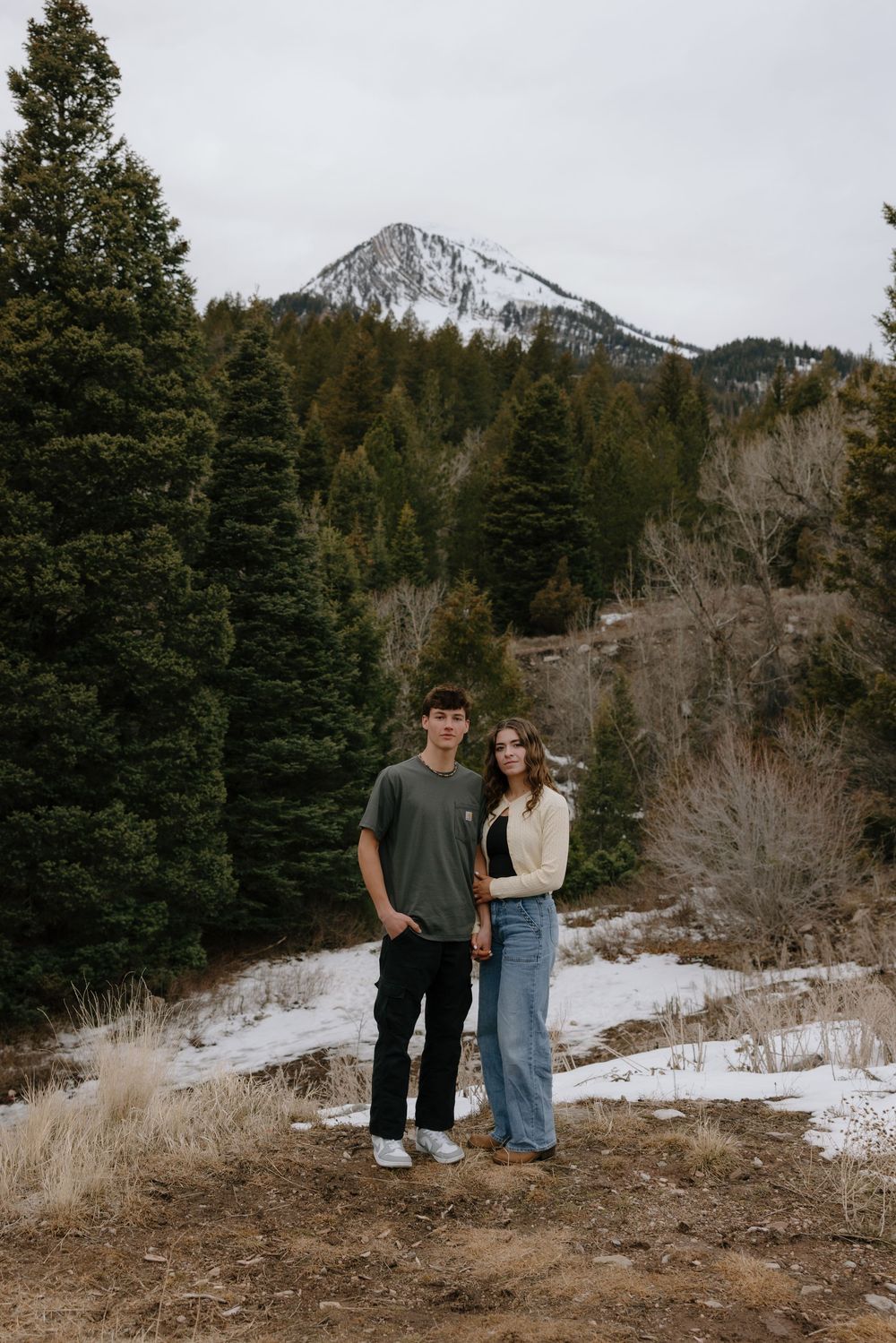 Young couple standing together in snowy mountain forest landscape with snow-capped peak backdrop.