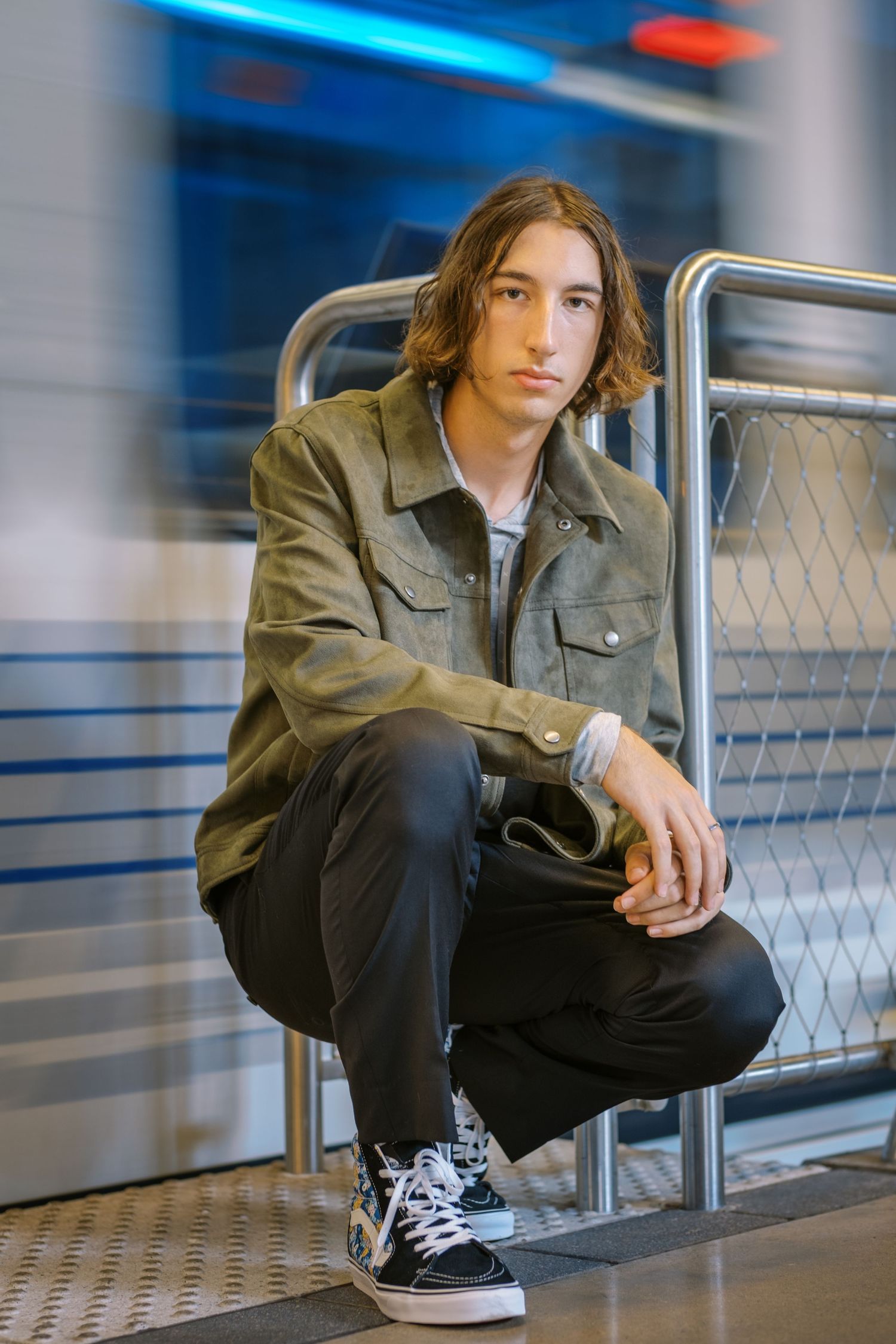 Young man in olive jacket and Vans sneakers crouching by metal railings in a modern transit station.