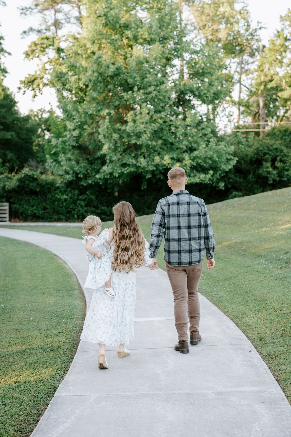 Three children walking hand-in-hand down a sidewalk path surrounded by lush green grass and trees.