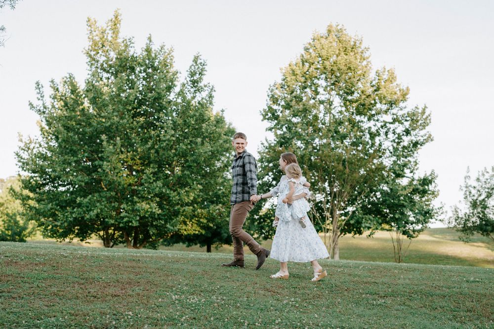 A couple walks hand-in-hand on a green lawn surrounded by lush trees, the woman in a white dress holding a baby.