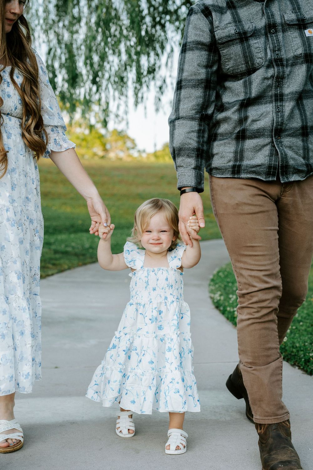 Toddler girl in floral dress walks holding parents hands on a park path outdoors.
